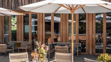 An outdoor dining area with wicker furniture and umbrellas, set against a wooden wall with large windows.