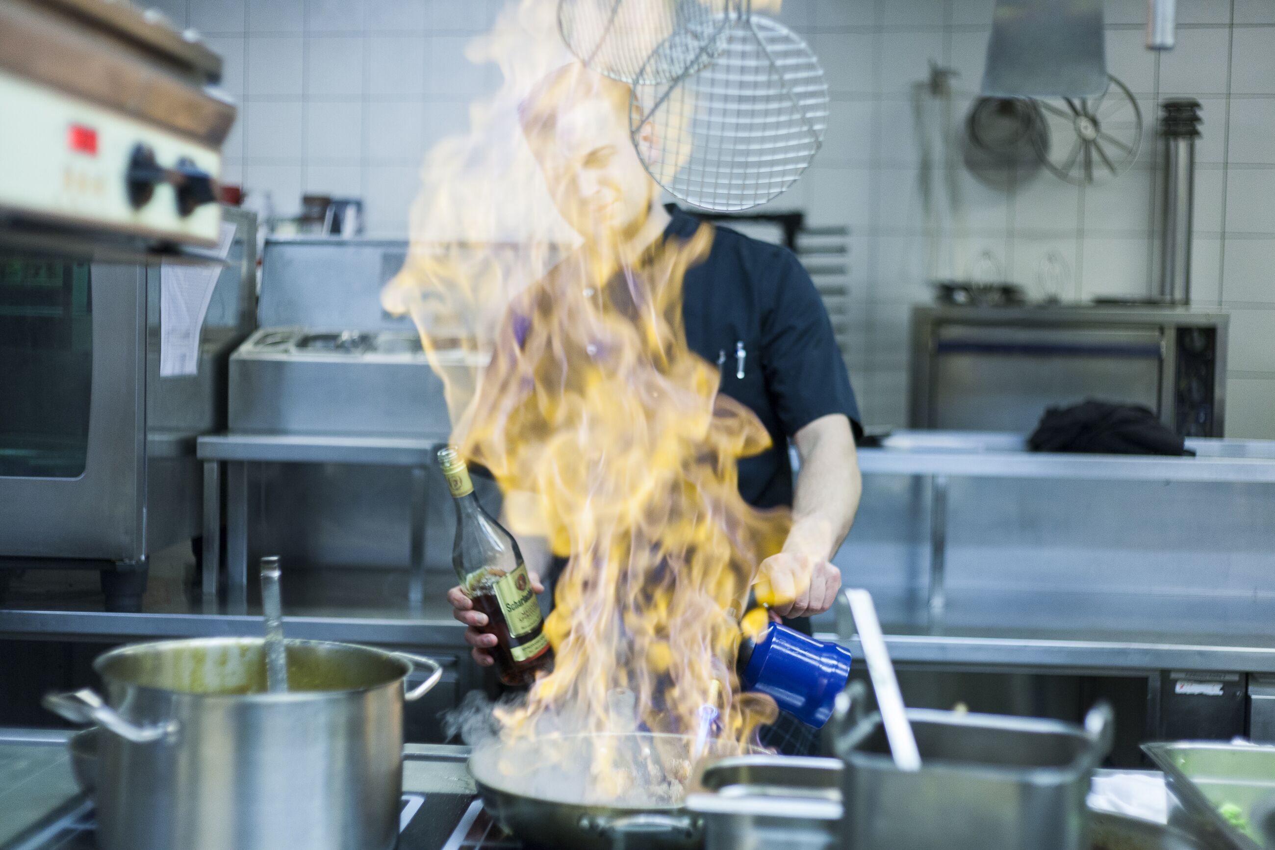 Una cocina con electrodomésticos de acero inoxidable, una olla en la hornilla y una persona sirviendo líquido en una sartén.