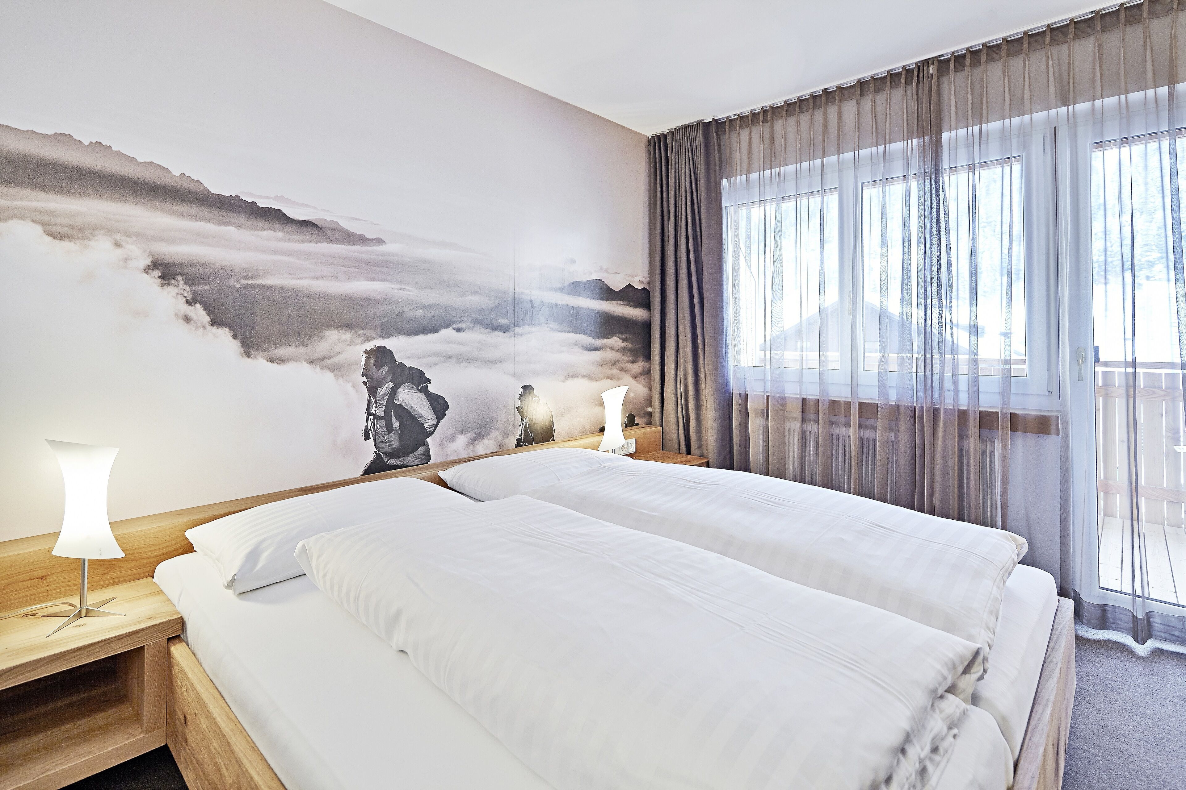 A hotel room with a large bed, a nightstand with a lamp, a wall mural, and a window with curtains.