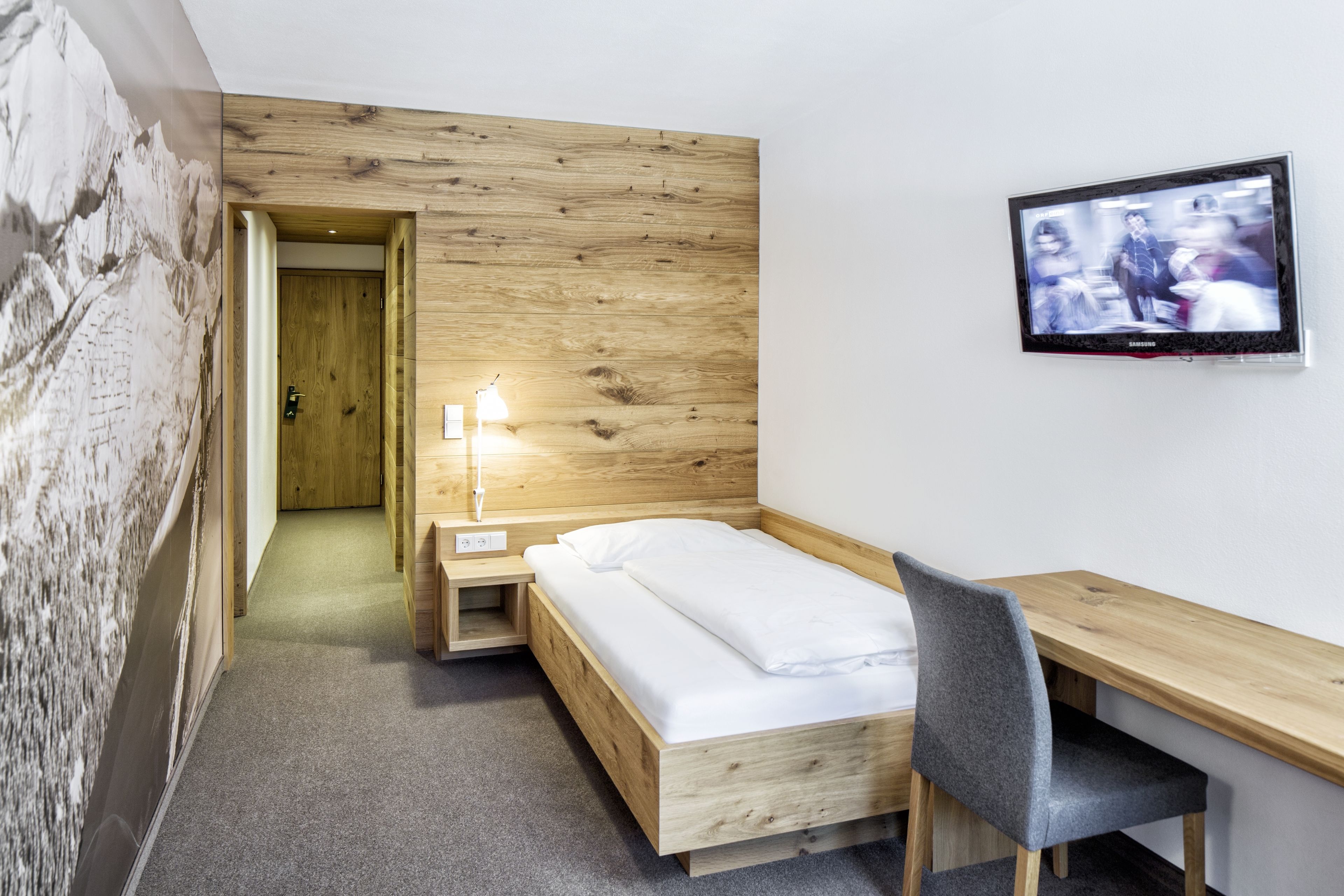 Una camera d'hotel moderna con una testiera in legno, un letto con lenzuola bianche, una scrivania con una sedia, una televisione a parete e una porta che dà su un'altra stanza.