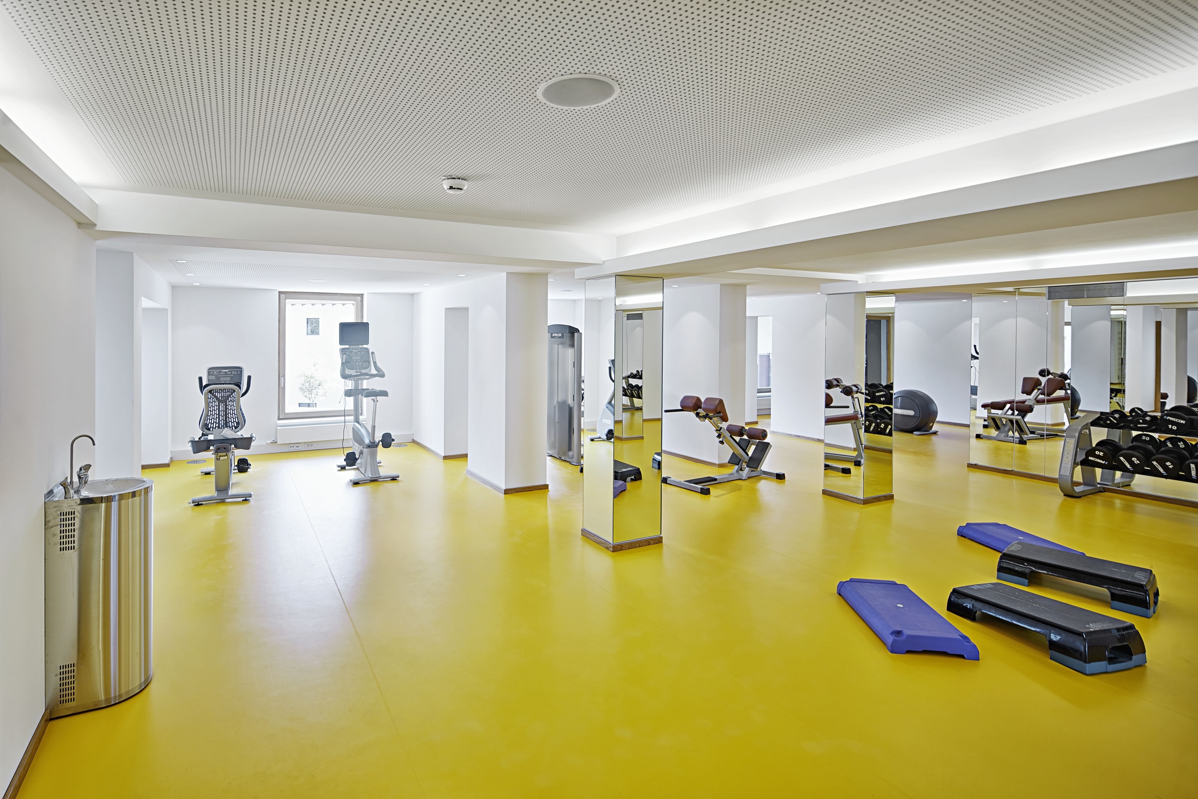 Un gimnasio moderno con piso amarillo, varios equipos de ejercicio y espejos.