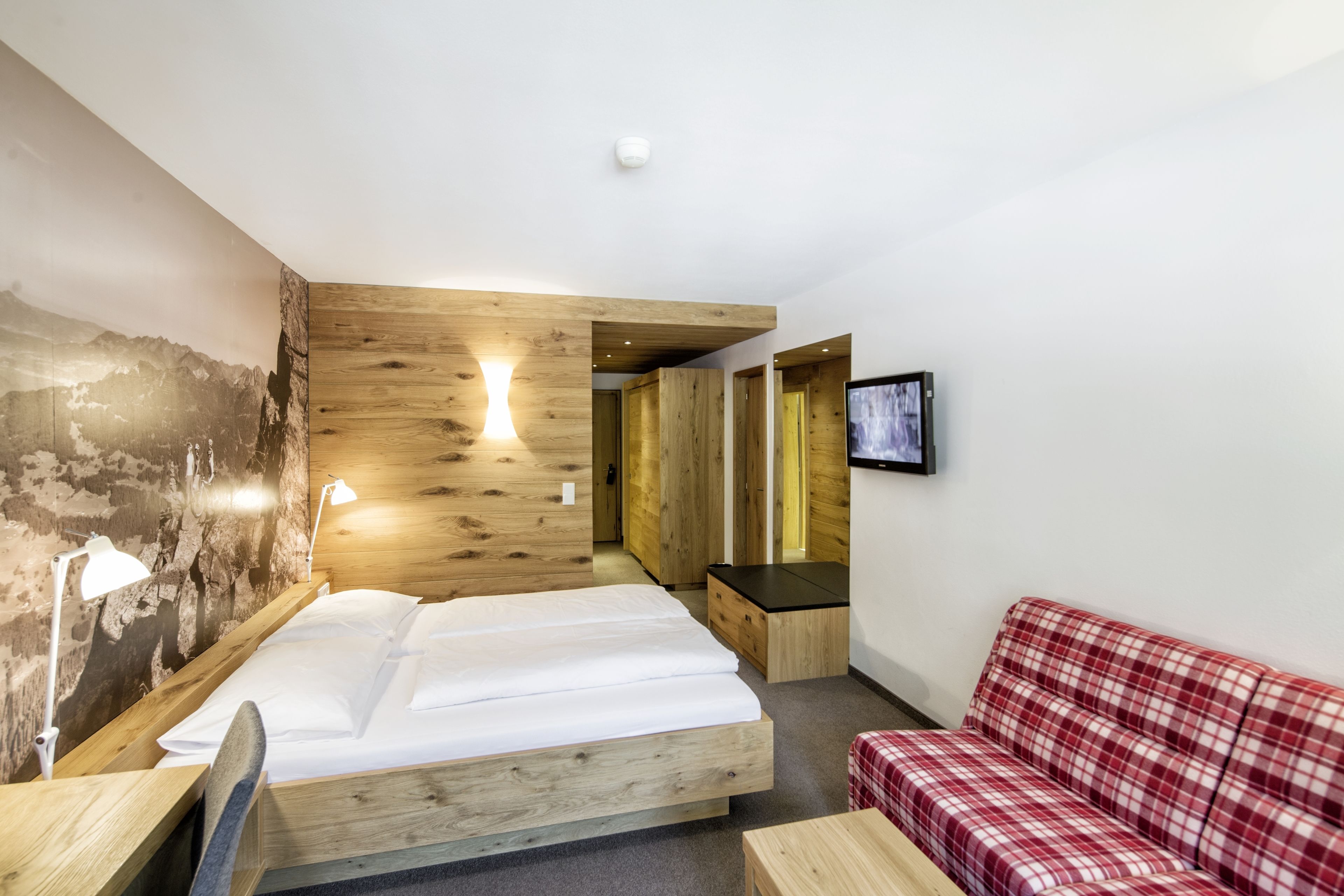 A modern hotel room with a wooden bed, a red plaid sofa, a mounted TV, and a large wall mural.