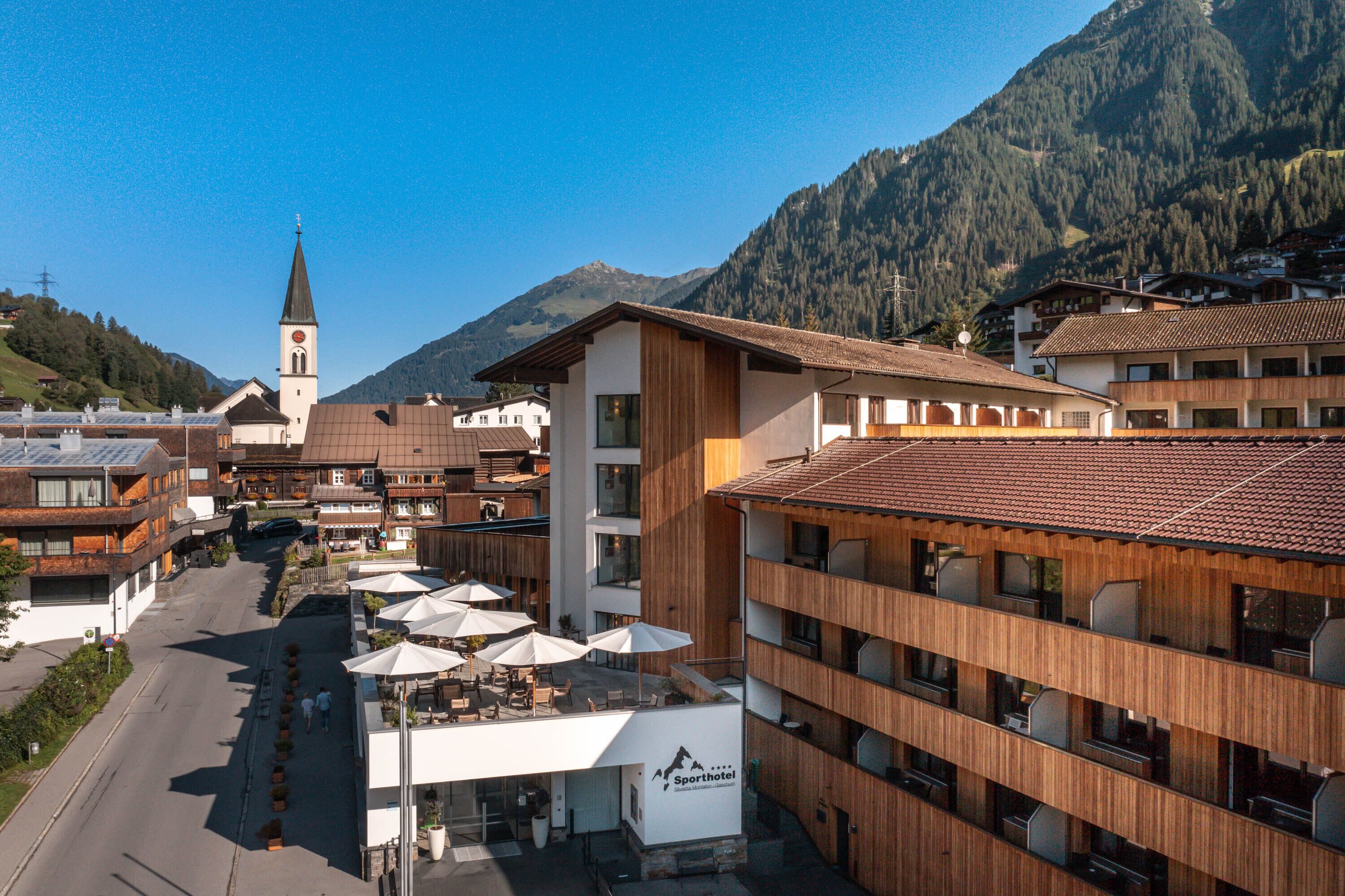Un hotel de diseño moderno, con una vista destacada de la montaña, una iglesia al fondo y mesas exteriores con sombrillas.