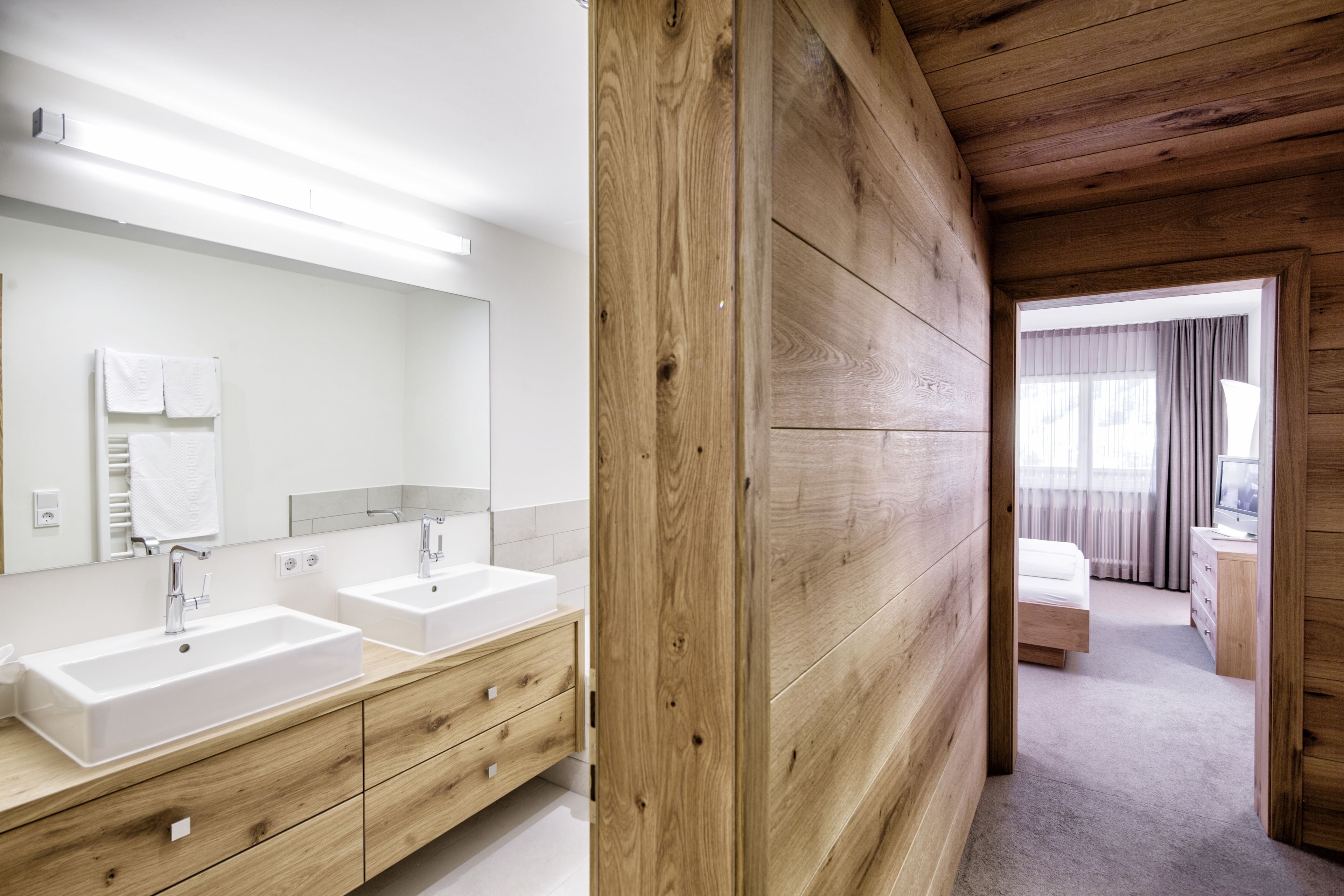 Un baño moderno con dos lavamanos, paneles de madera y una habitación visible con cama y cómoda.