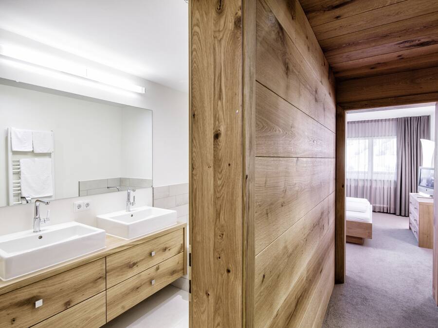 A modern bathroom with two sinks, wooden paneling, and a visible bedroom with a bed and dresser.