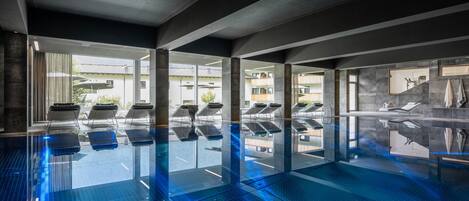 Une piscine intérieure moderne avec des parois en verre transparent, des chaises longues et une vue sur les bâtiments environnants.