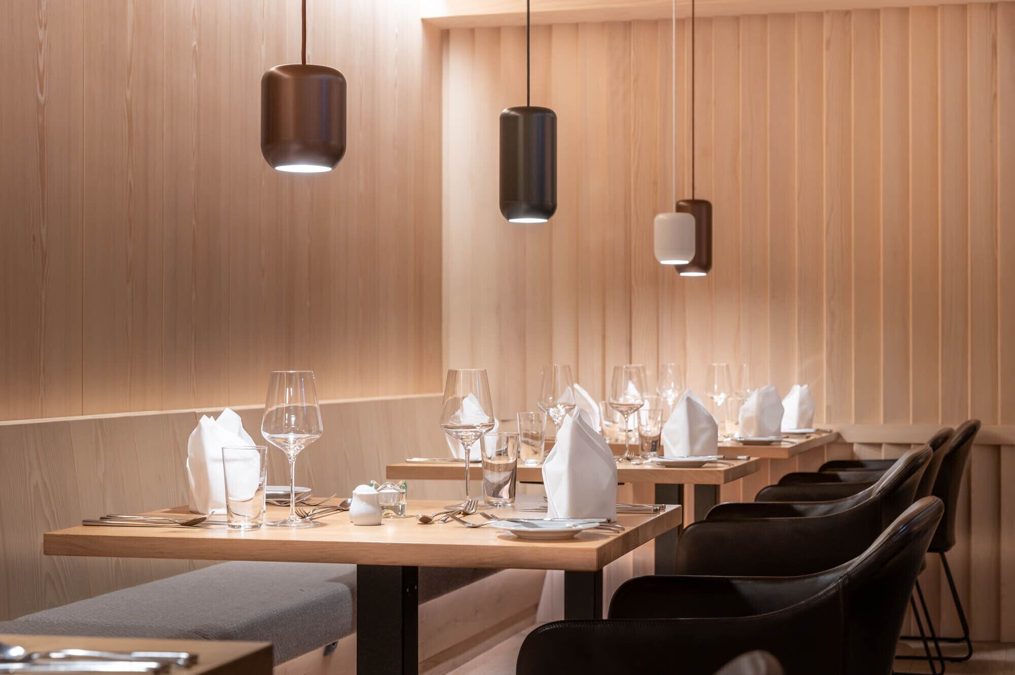 A modern restaurant with wooden panel walls, black leather chairs, and neatly set dining tables with wine glasses and napkins.