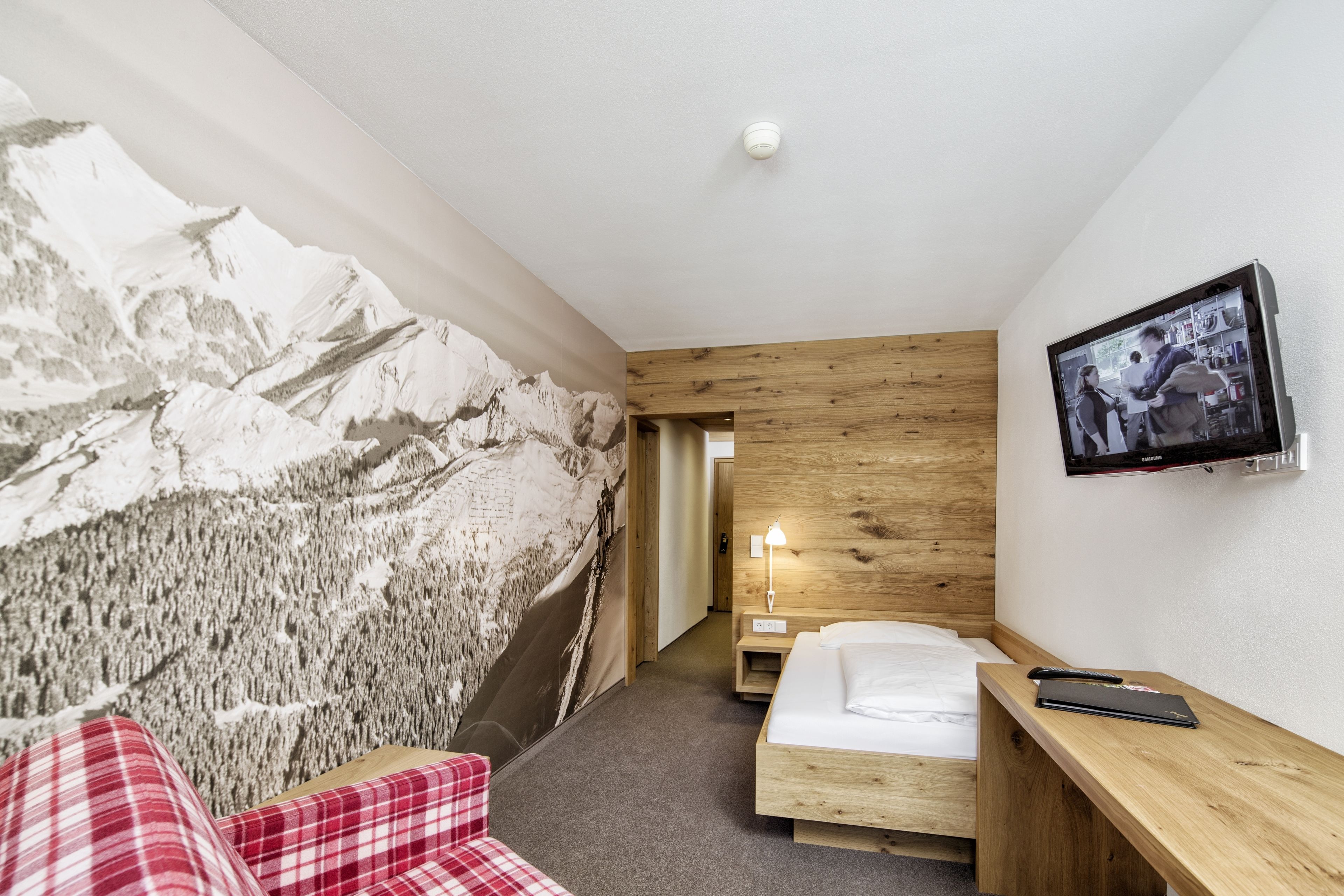 A hotel room with a bed, a wooden desk, a mounted TV, and a wall mural of mountains.
