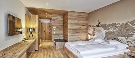 A modern bedroom with a wooden floor, a bed with white bedding, a desk with a lamp, and a large mirror.