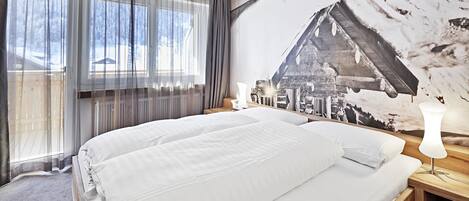 Un dormitorio con una cama grande, una mesita de noche con lámpara, una ventana con cortinas traslúcidas y un mural en la pared que muestra un paisaje nevado de montaña.