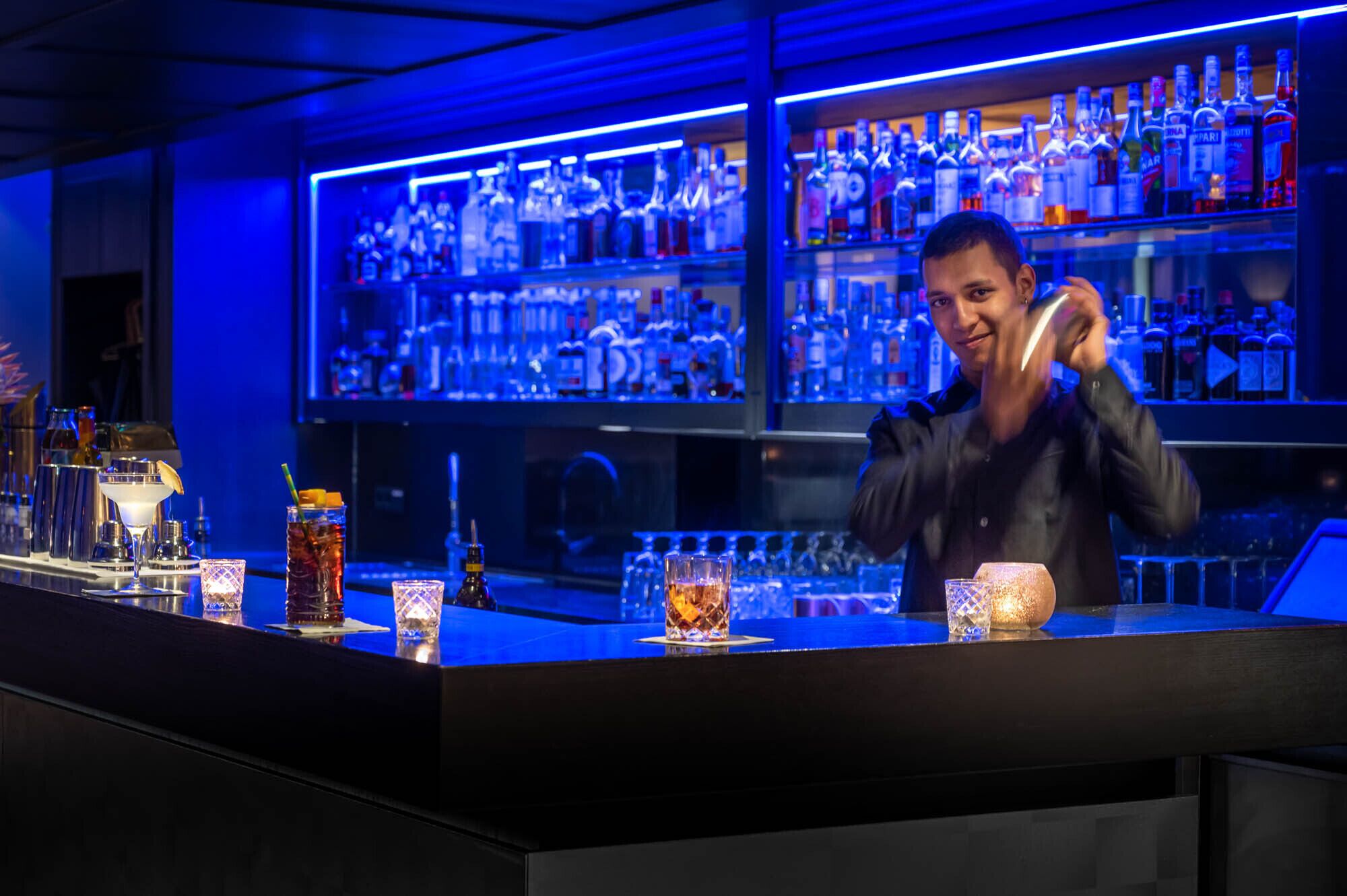 Un bar con un cantinero preparando bebidas, estantes con botellas de licores y un interior bien iluminado.
