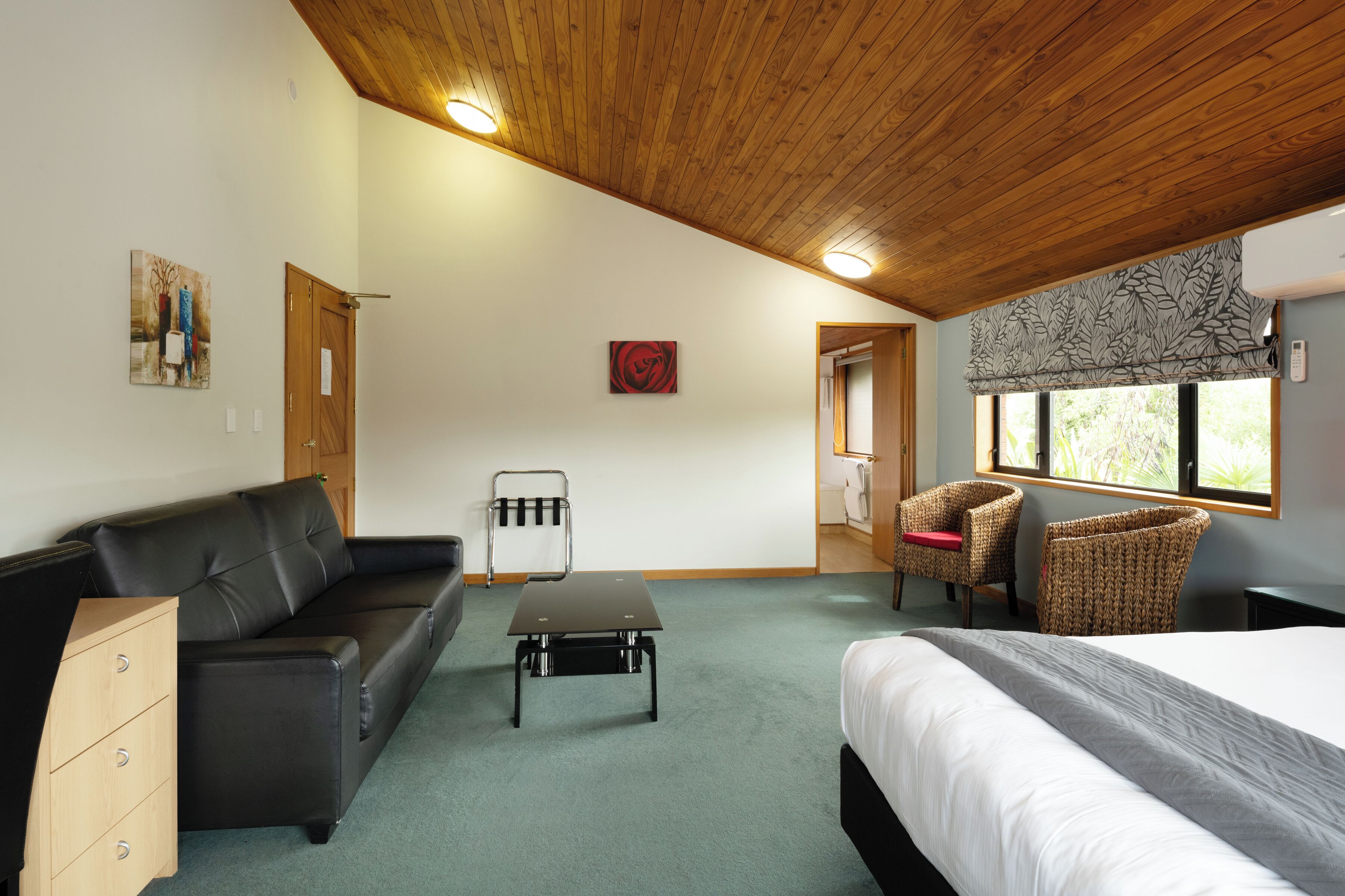 Una stanza con soffitto in legno, un divano in pelle nera, un letto con biancheria bianca, un tavolino, due poltrone in vimini e una finestra con vista sul verde.