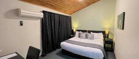 A hotel room with a wooden ceiling, a bed with white bedding and patterned pillows, a desk with a chair, and a wall-mounted air conditioner.