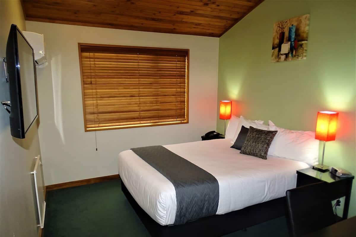 Ein Hotelzimmer mit Bett, Nachttischlampen, Fernseher, Fenster mit Jalousien und einem Bild an der Wand.