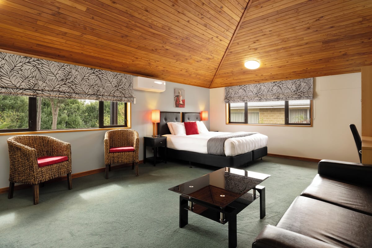 Ein Schlafzimmer mit einem Bett, zwei Korbstühlen mit roten Kissen, einem Holztisch und Blick auf Bäume durch die Fenster.