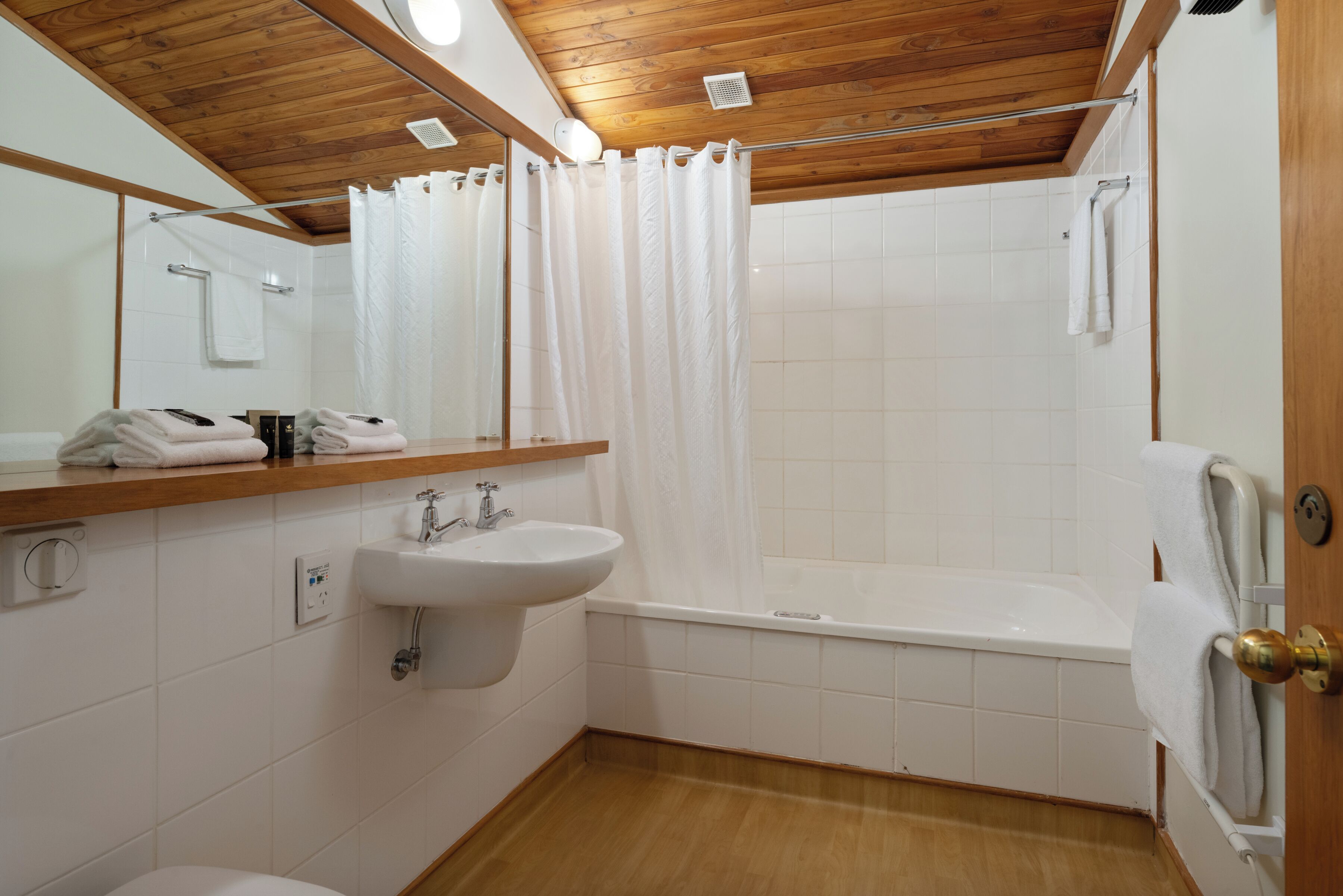 A bathroom with a wooden ceiling, white tiles, a sink, a bathtub, and a mirror.
