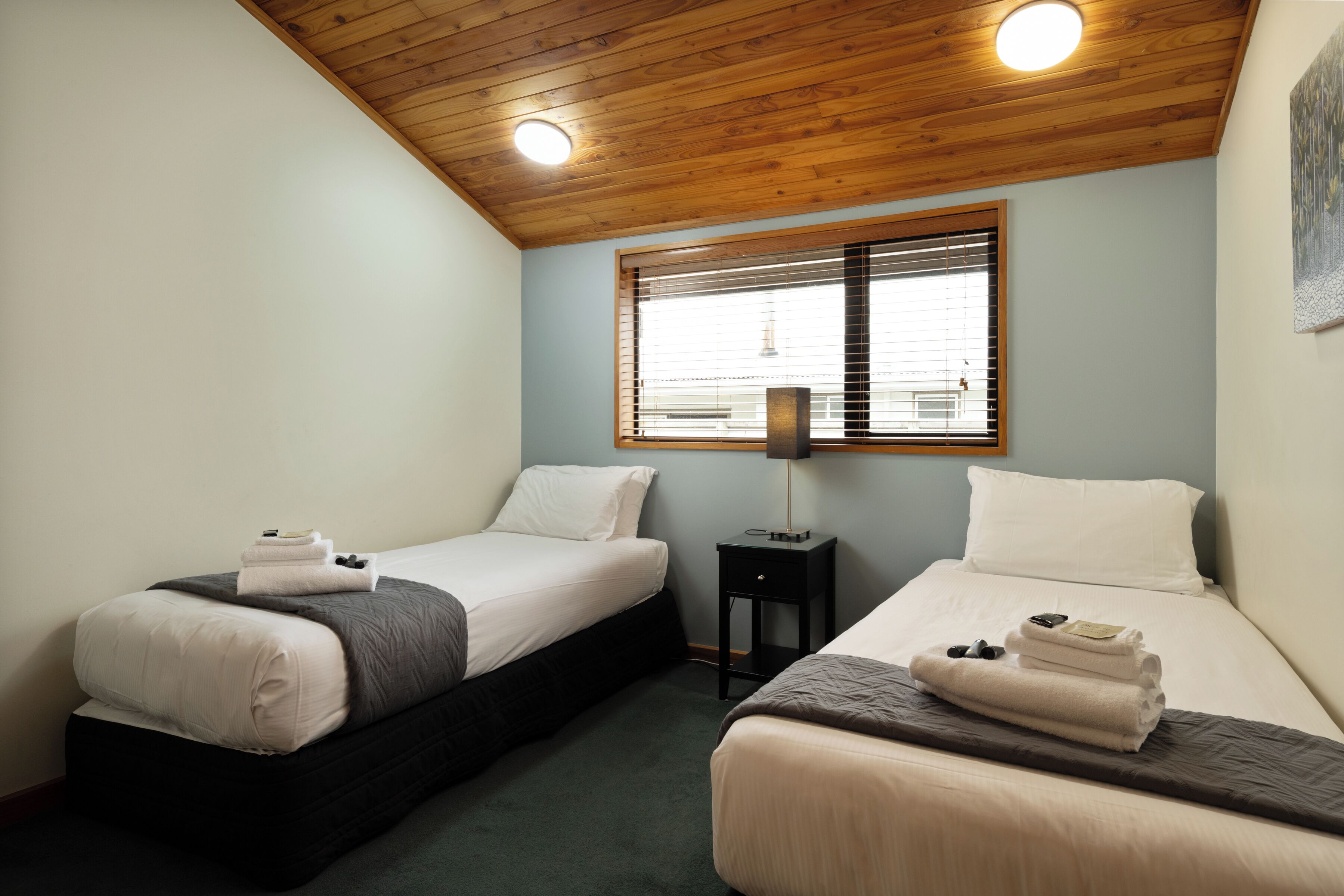 A room with two beds, a window with blinds, a small table with a lamp, and a ceiling light.