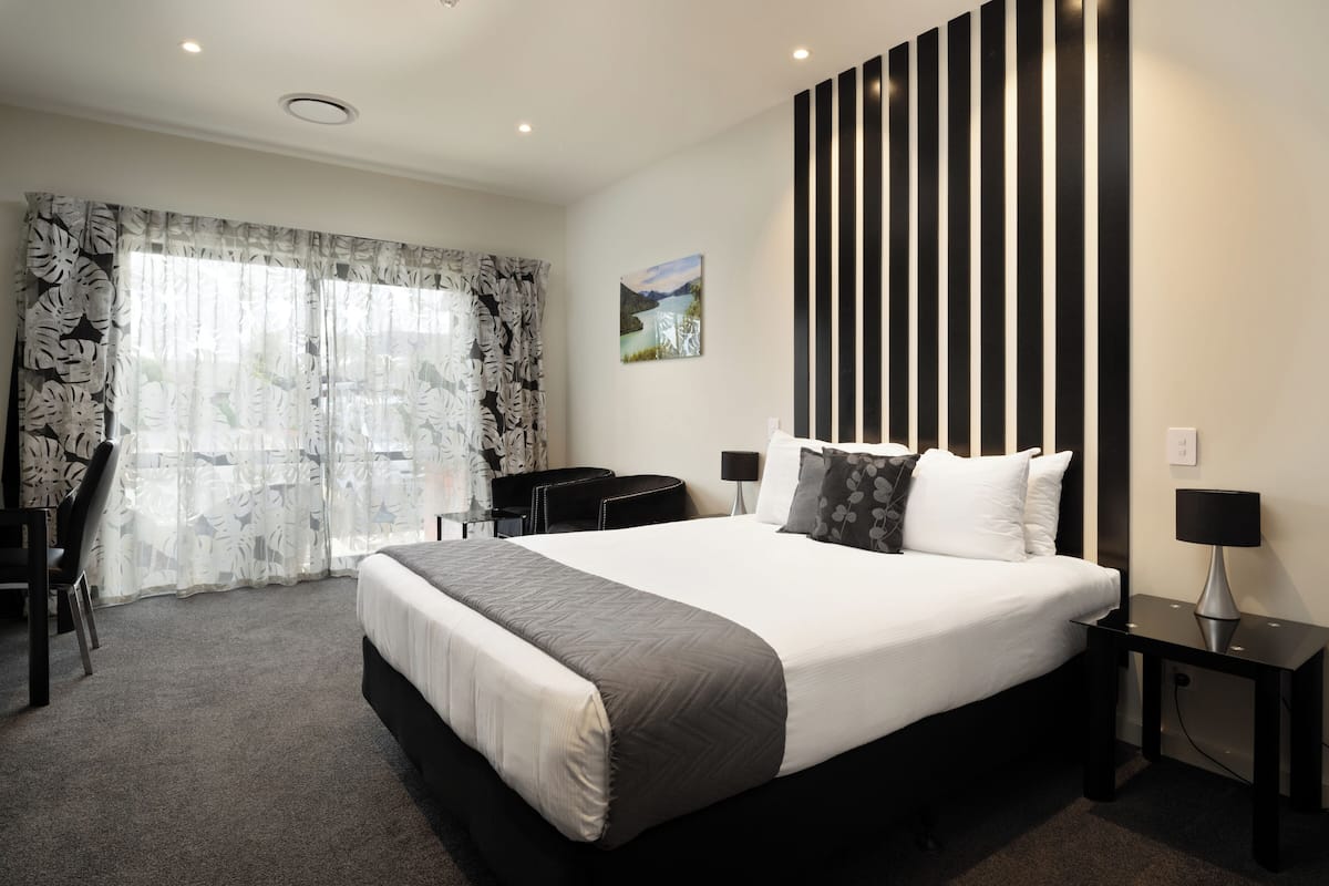 Ein modernes Hotelzimmer mit einem großen Bett, einem Schreibtisch und einem Sessel. Ein Fenster mit Vorhängen, ein Bild an der Wand und eine schwarze, vertikale Leuchte sind zu sehen.