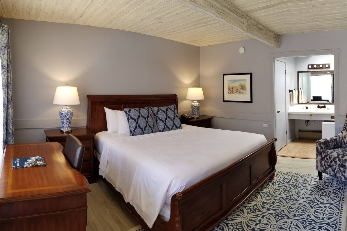 A hotel room with a wooden bed, two bedside tables with lamps, a patterned rug, a chair, and a bathroom visible through an open door.
