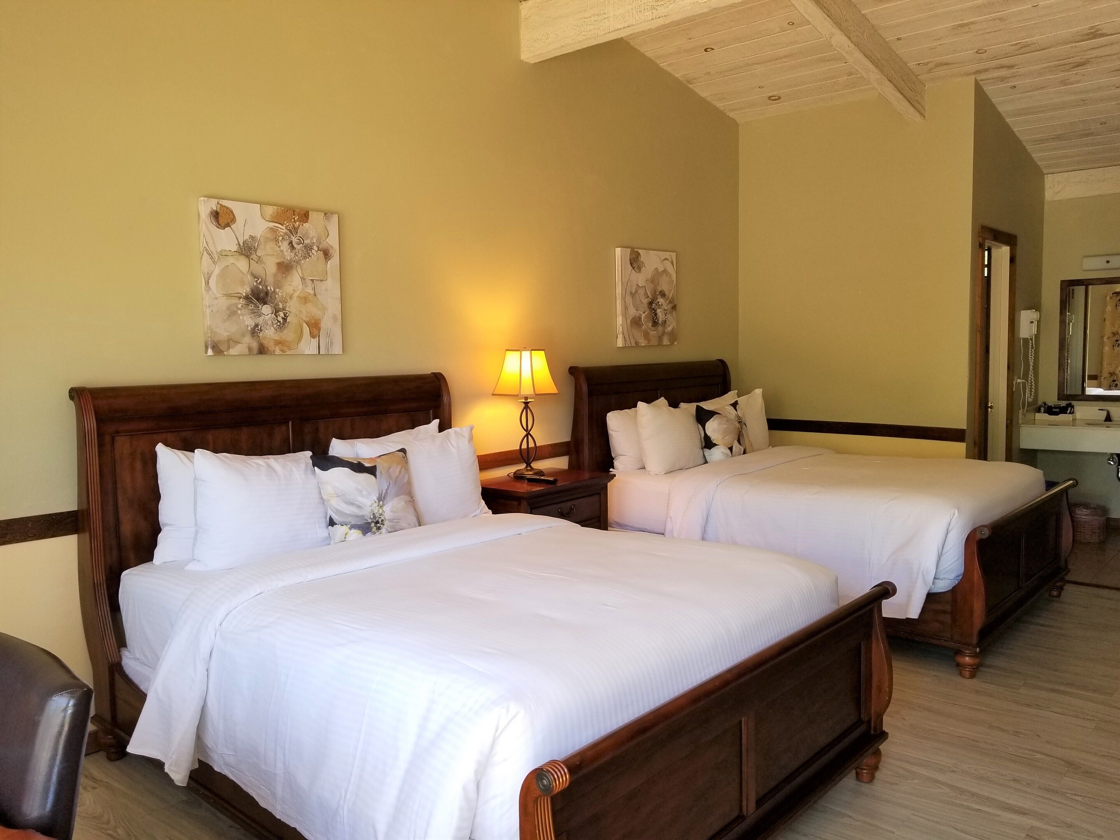 Habitación de hotel con dos camas, cabeceros de madera, ropa de cama blanca, una lámpara de noche y obras de arte en las paredes.