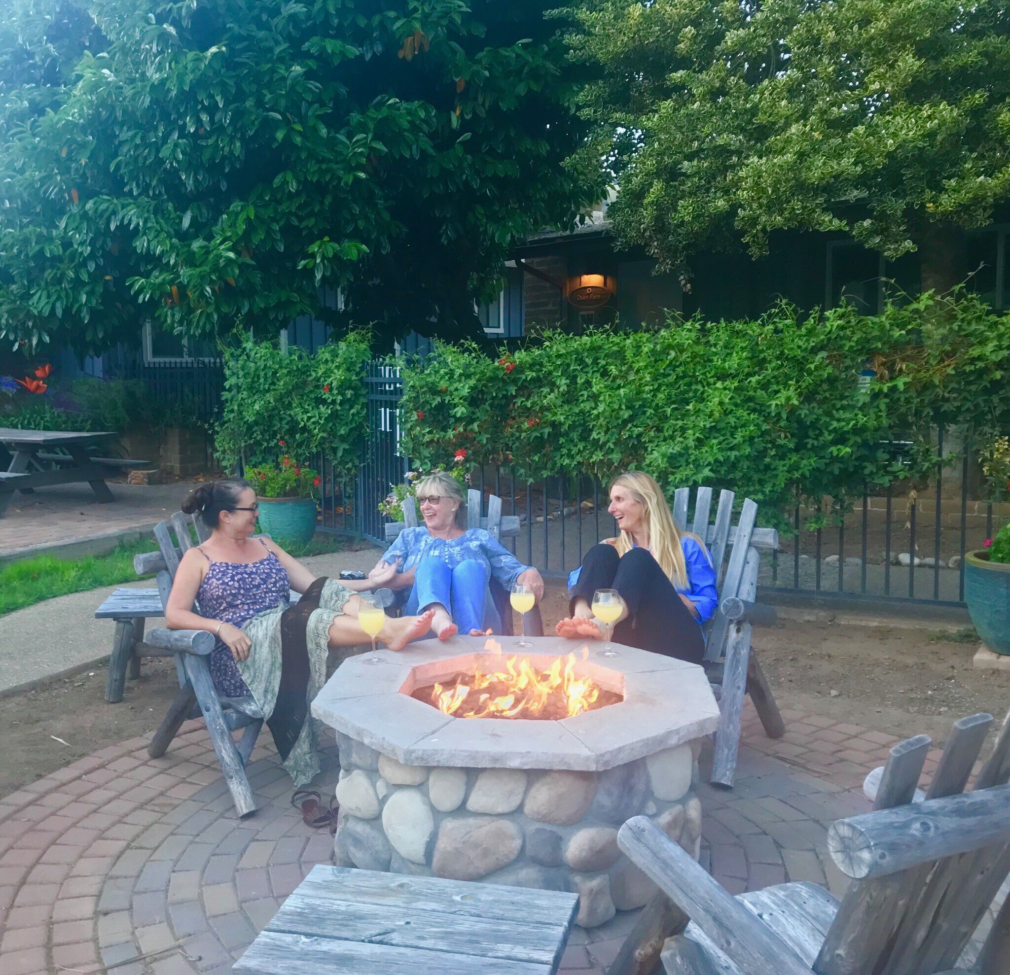 Three people seated around a stone fire pit with drinks, surrounded by greenery and a building in the background.
