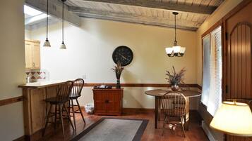 Une cuisine avec des meubles en bois, une table à manger avec des chaises, une horloge, des luminaires suspendus et une fenêtre avec des stores.