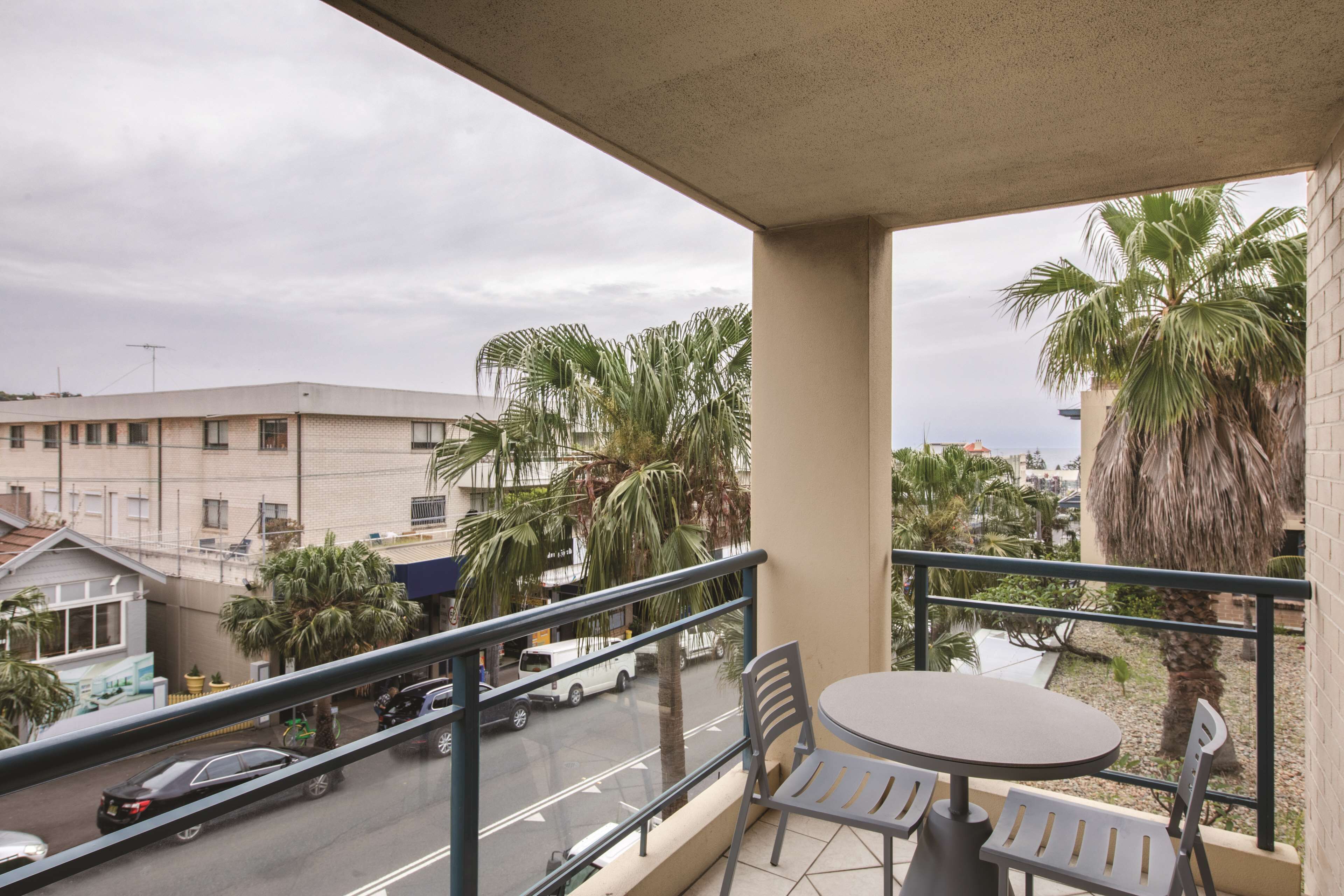 Un balcon avec une table et des chaises, donnant sur une rue bordée de palmiers et sur laquelle on aperçoit un bâtiment au loin.