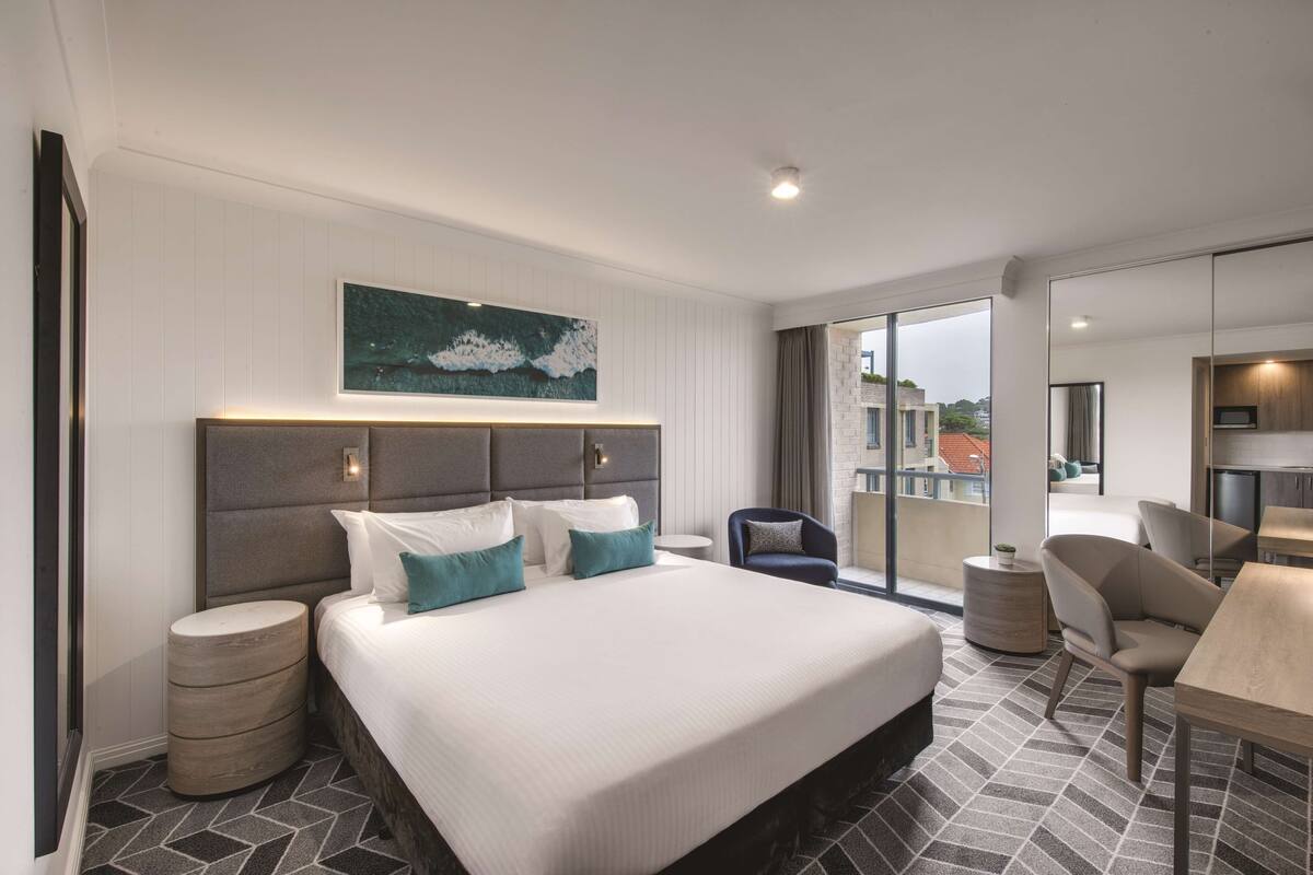 Een moderne hotelkamer met een groot bed, een zithoek met een tafel, en uitzicht op de stad door grote ramen.