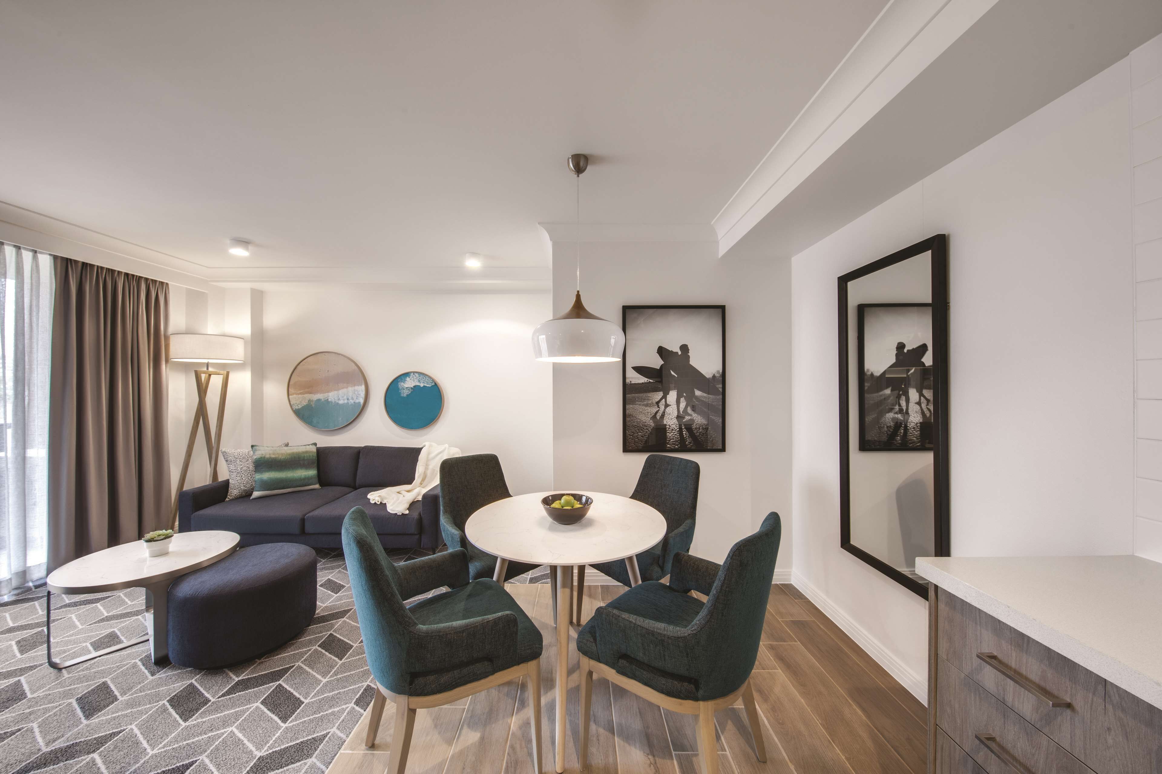 A modern living room with a sofa, round table, and chairs, a dining area with a bowl of fruit, and artwork on the walls.
