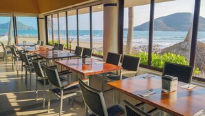 Un restaurante con amplios ventanales que ofrecen vistas al océano, mesas y sillas dispuestas para comer, y una decoración tropical con plantas.