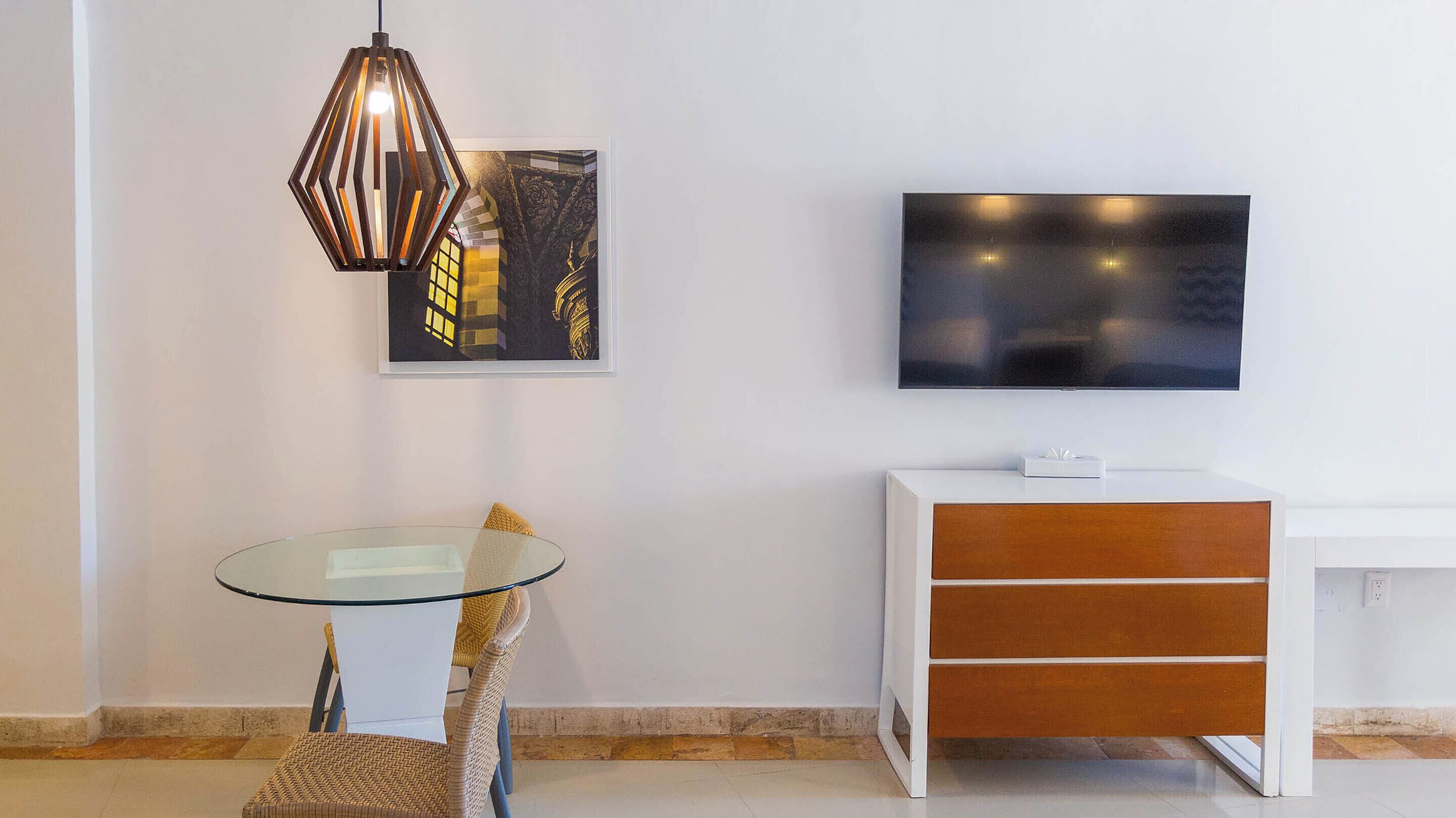 A modern living room with a glass table, wicker chair, a decorative hanging light, a flat-screen TV, and a white cabinet with drawers.