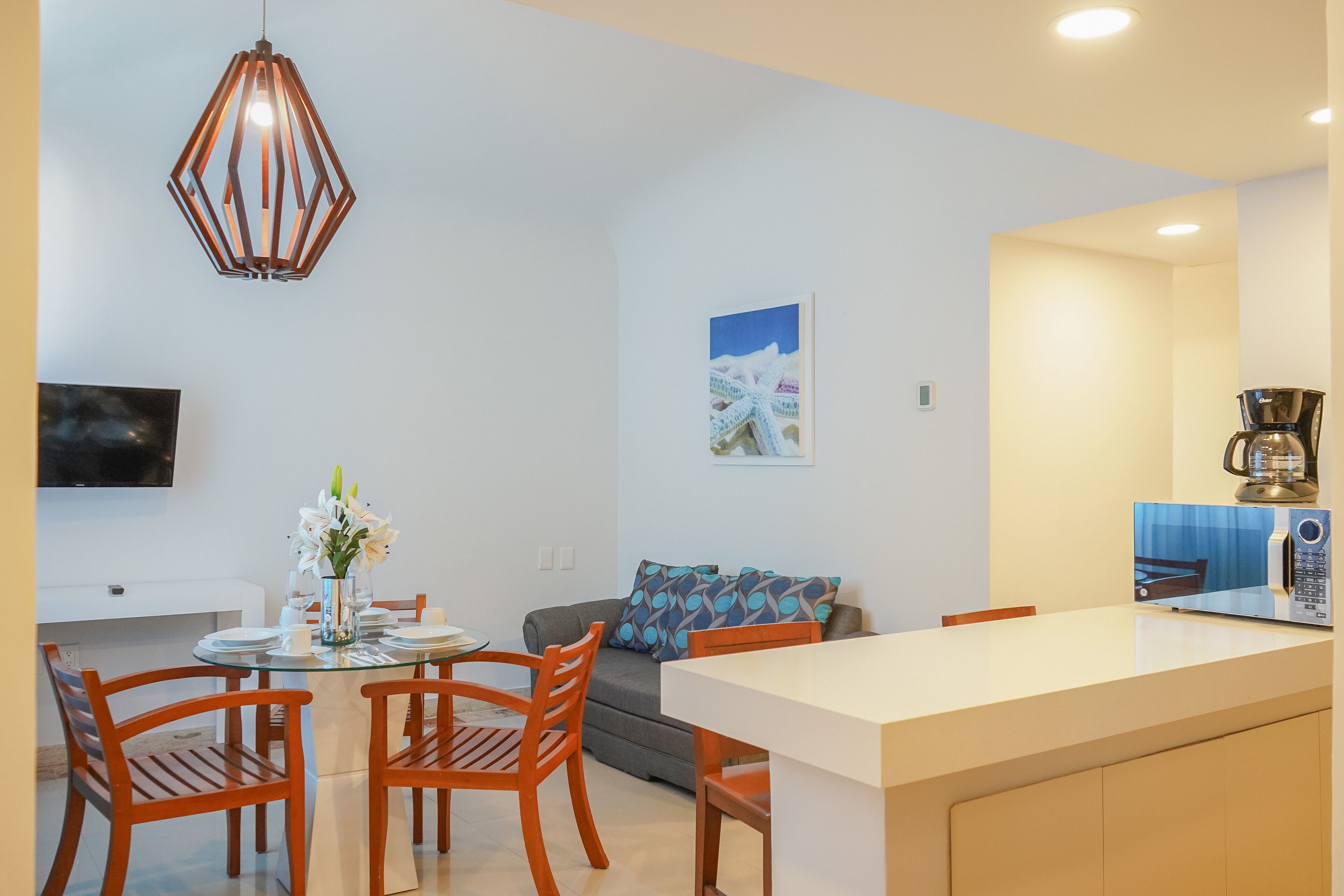 A modern kitchen with a built-in coffee maker, a dining area with wooden chairs, and a gray sofa with patterned cushions.