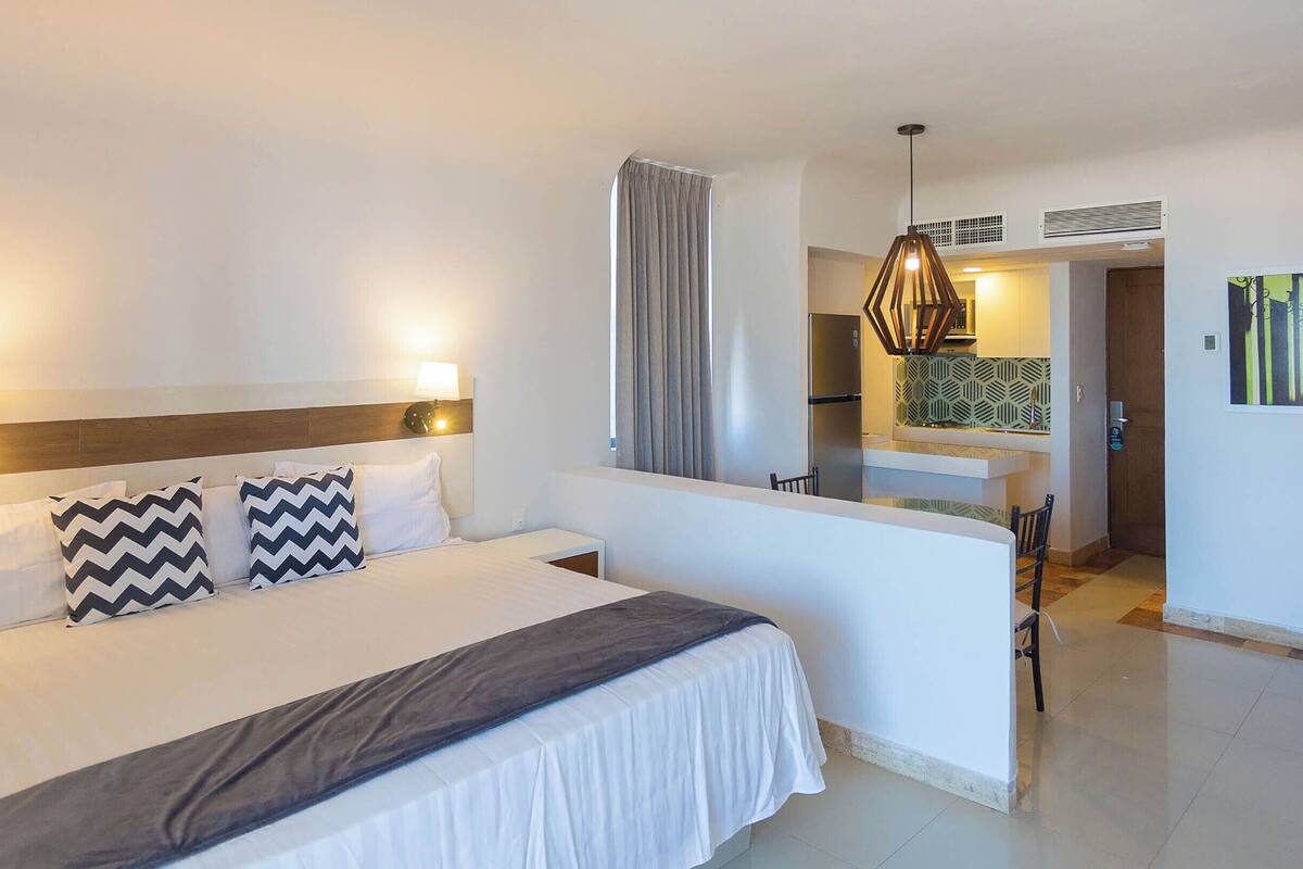 A hotel room with a bed, bedside lamps, a window with curtains, a kitchenette, and a hallway leading to another room.