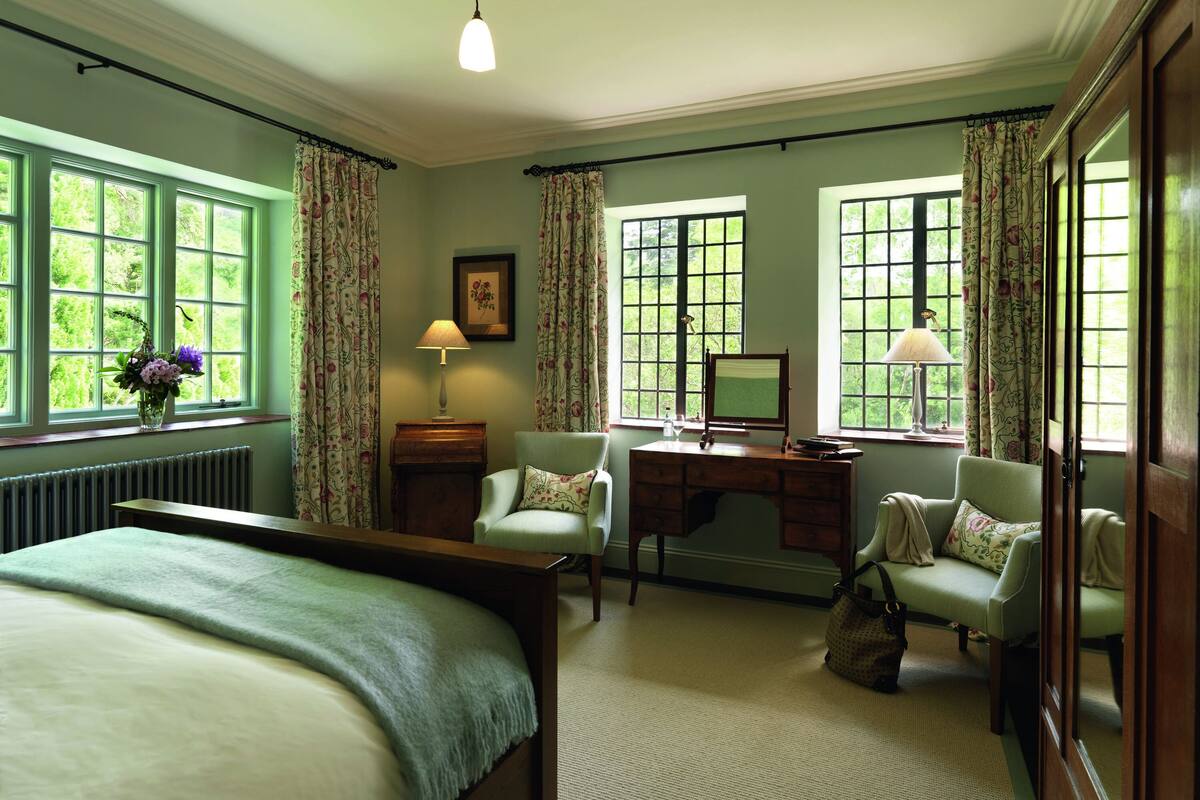 A room with a bed, two chairs, a desk, a radiator, and large windows with curtains.
