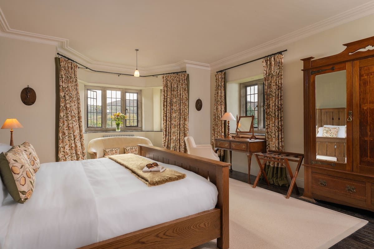 A bedroom with a large bed, a wooden dresser, a desk, and a chair. There are curtains on the windows, and a lamp is on the nightstand.