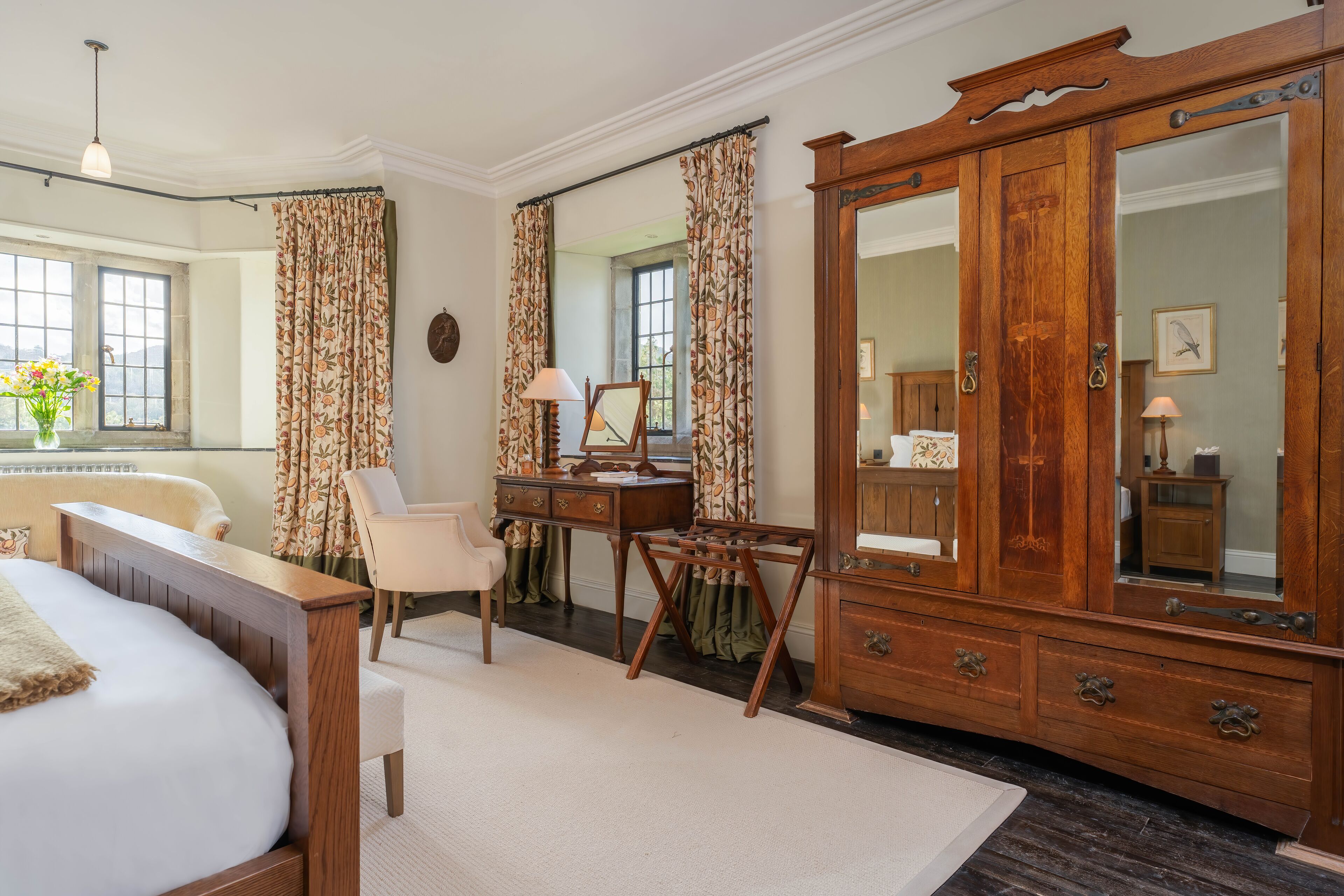 A bedroom with a large wooden wardrobe, a desk with a lamp, a chair, and a bed with a white comforter.