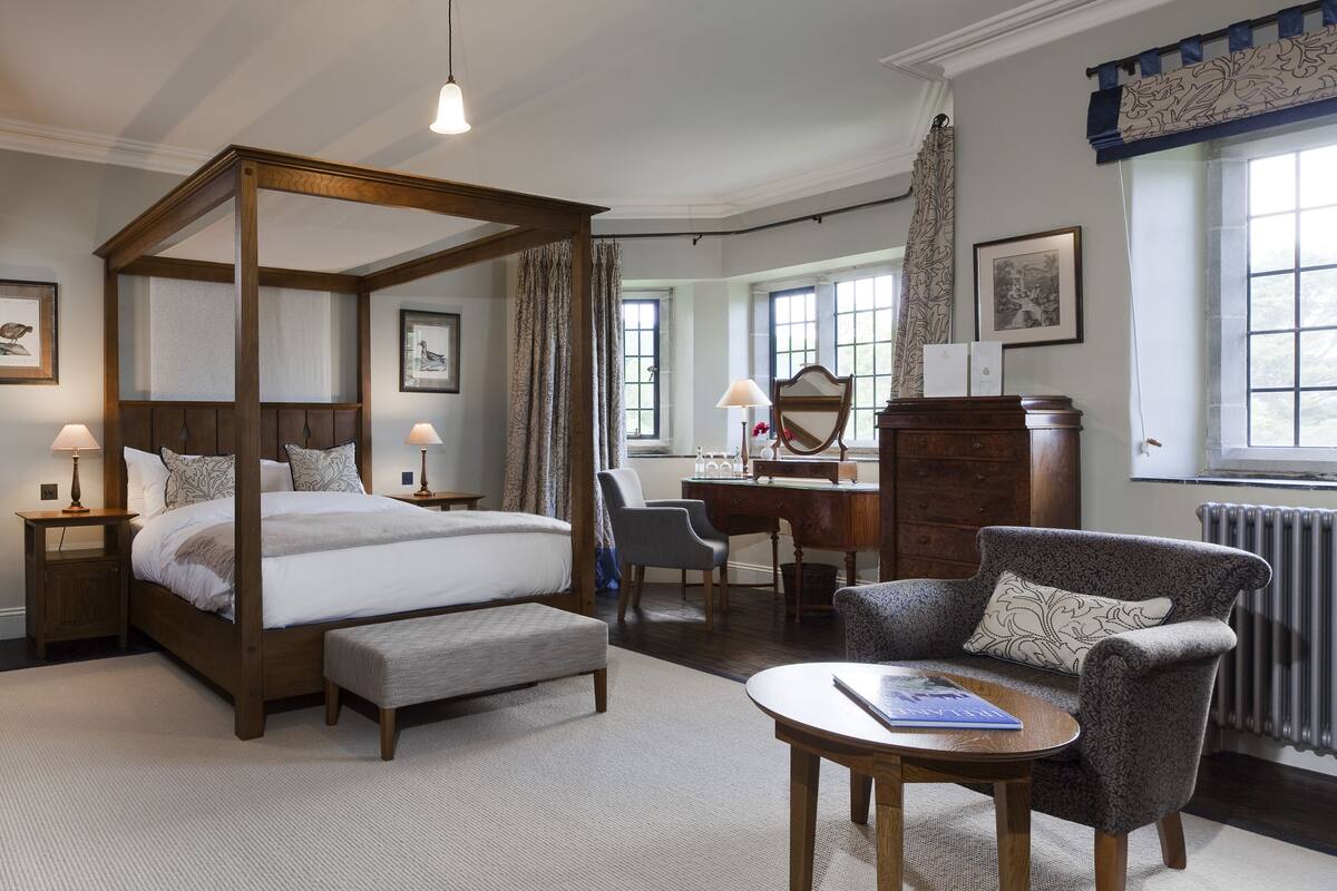 A four-poster bed with a wooden frame, a desk with a mirror, a chair, and a small table with a book.