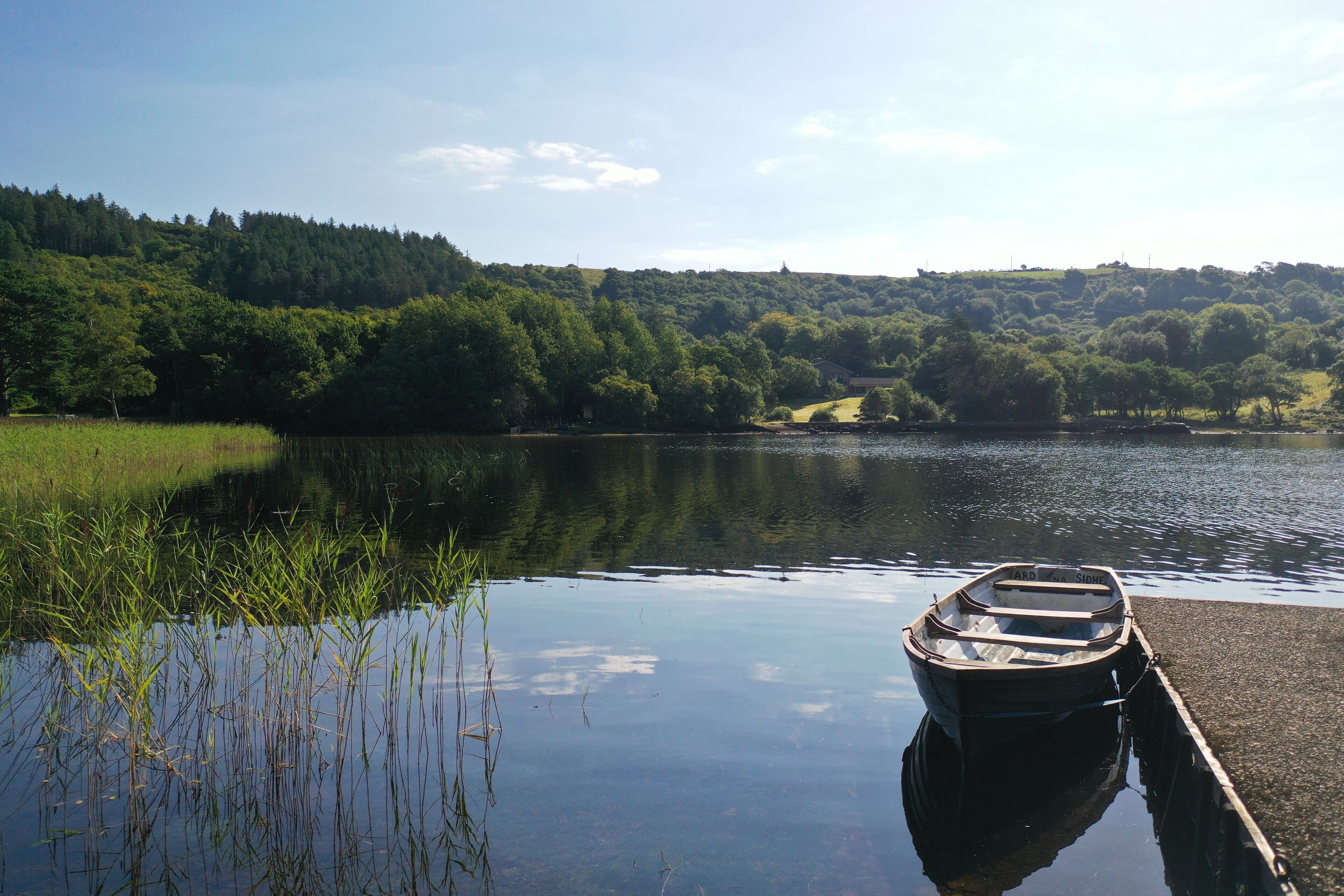 A calm lake with a wooden boat docked at the shore, surrounded by lush greenery and hills in the background.