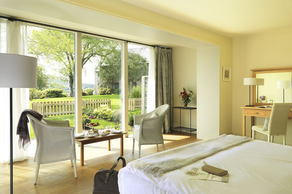 A hotel room with a large bed, a desk, a chair, a table with a tray of food, and a view of a garden through the window.