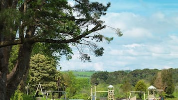 Children can play on swings, slides, and climbing structures in a safe, well-maintained park setting surrounded by lush greenery and trees.