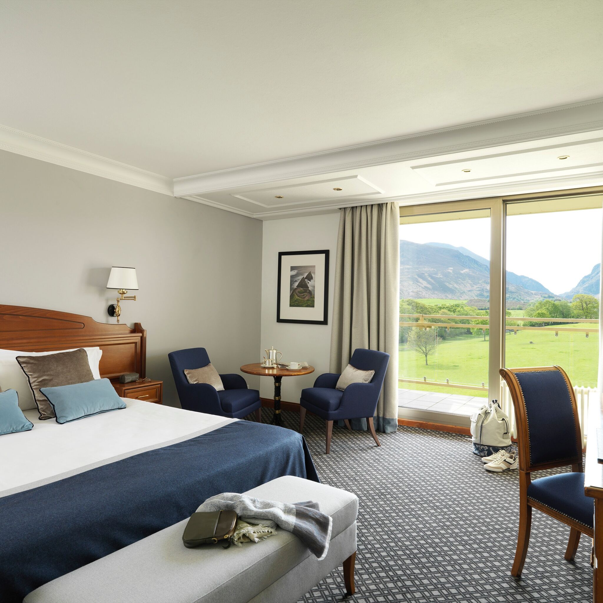 A hotel room with a large bed, two armchairs, a small table, and a view of mountains through large windows.
