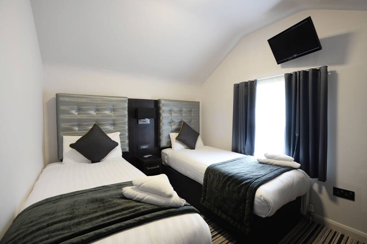 Ein Hotelzimmer mit zwei Betten, einem an der Wand montierten Flachbildfernseher und einem Fenster mit Vorhängen.