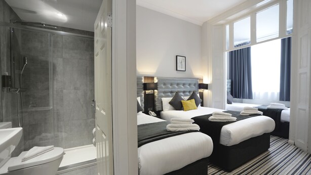 Ein modernes Hotelzimmer mit Dusche, zwei Betten, einem großen Fenster und Blick auf die Stadt.