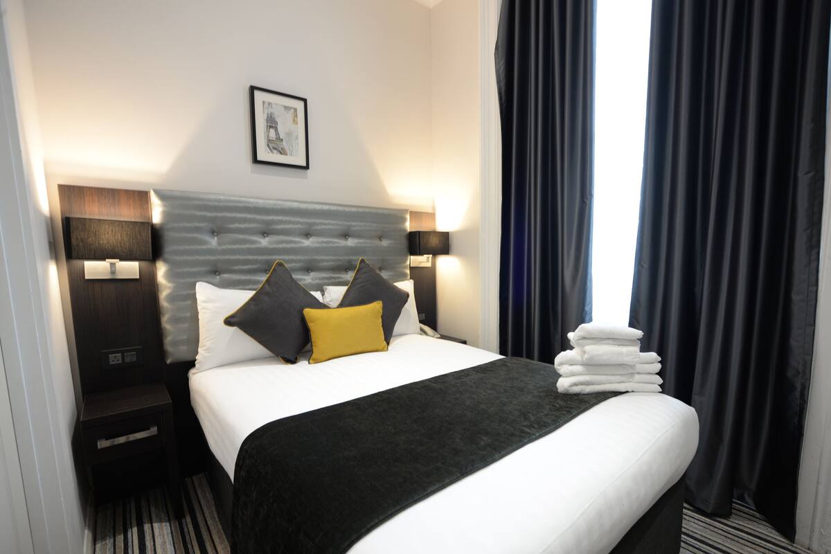 Ein Hotelzimmer mit einem ordentlich bezogenen Bett, einem Vorhang mit einem Bild, einem Nachttisch und einem Stapel Handtücher.