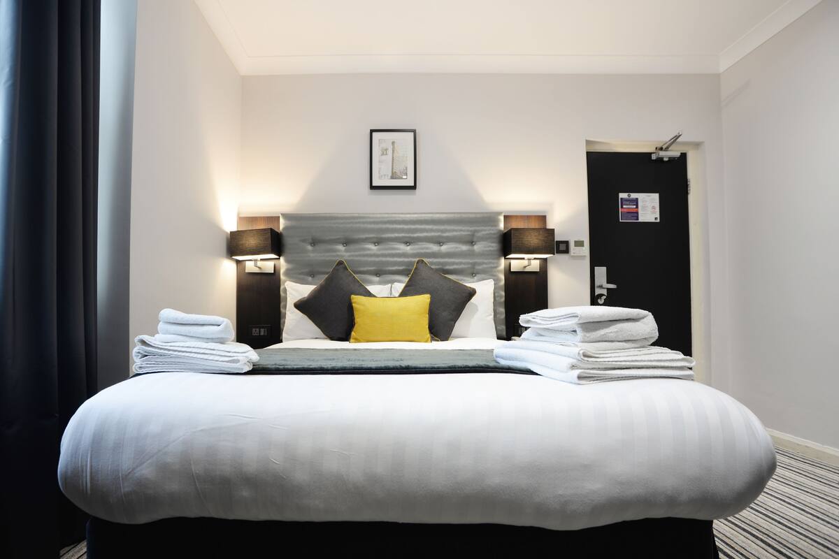 Ein Hotelzimmer mit einem großen Bett, zwei Nachttischlampen, einem gerahmten Bild an der Wand und einer Tür mit einem Schild.