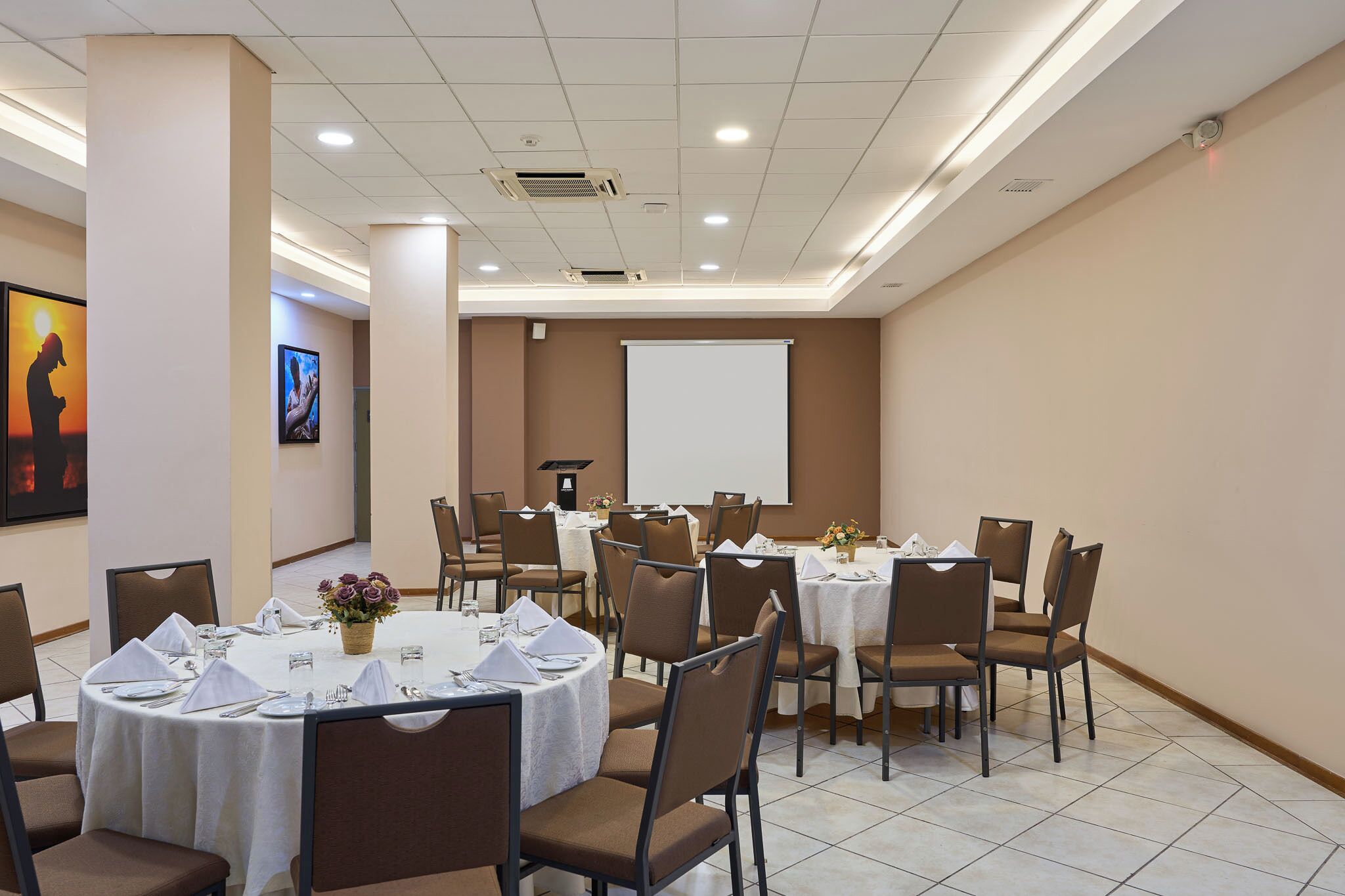 A conference room with multiple tables and chairs, a projector screen, and a large painting on the wall.