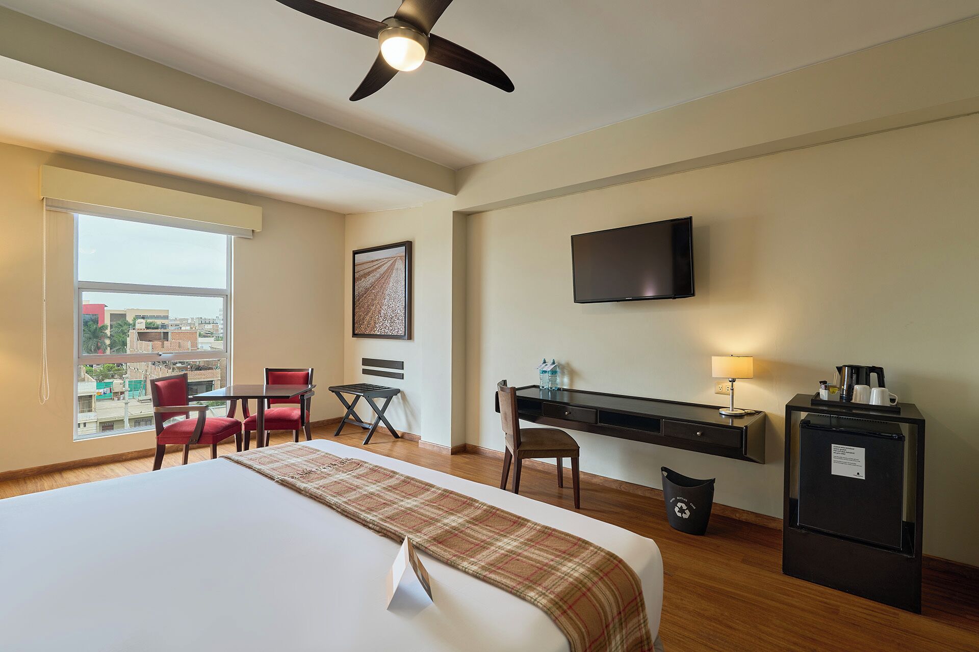 A hotel room with a bed, desk, chairs, a television, and a balcony with a view of buildings.