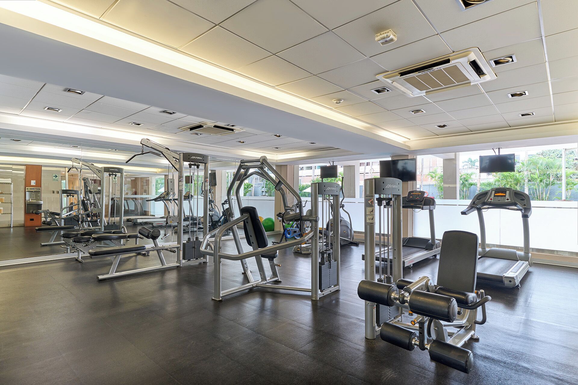 Een sportschool met diverse fitnessapparaten, grote ramen en een spiegelwand.