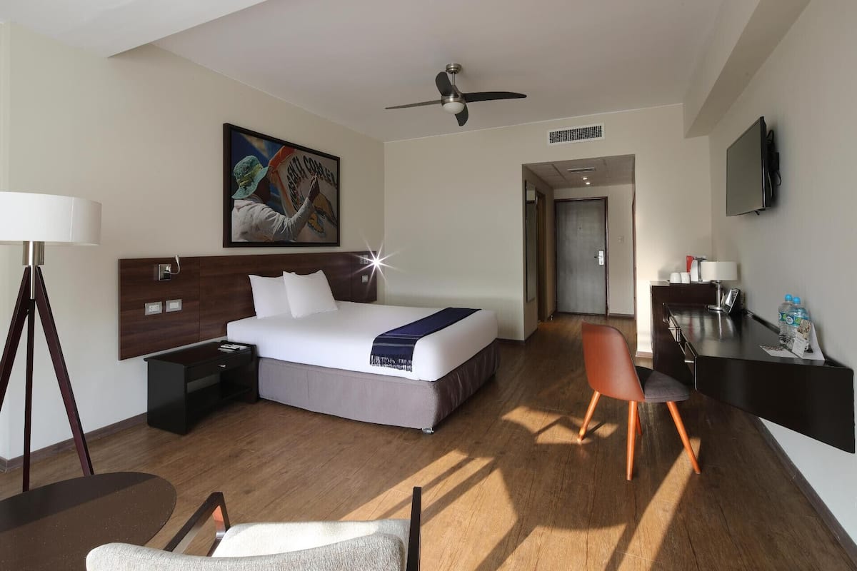 A hotel room with a bed, desk, chair, lamp, ceiling fan, artwork, and a television.