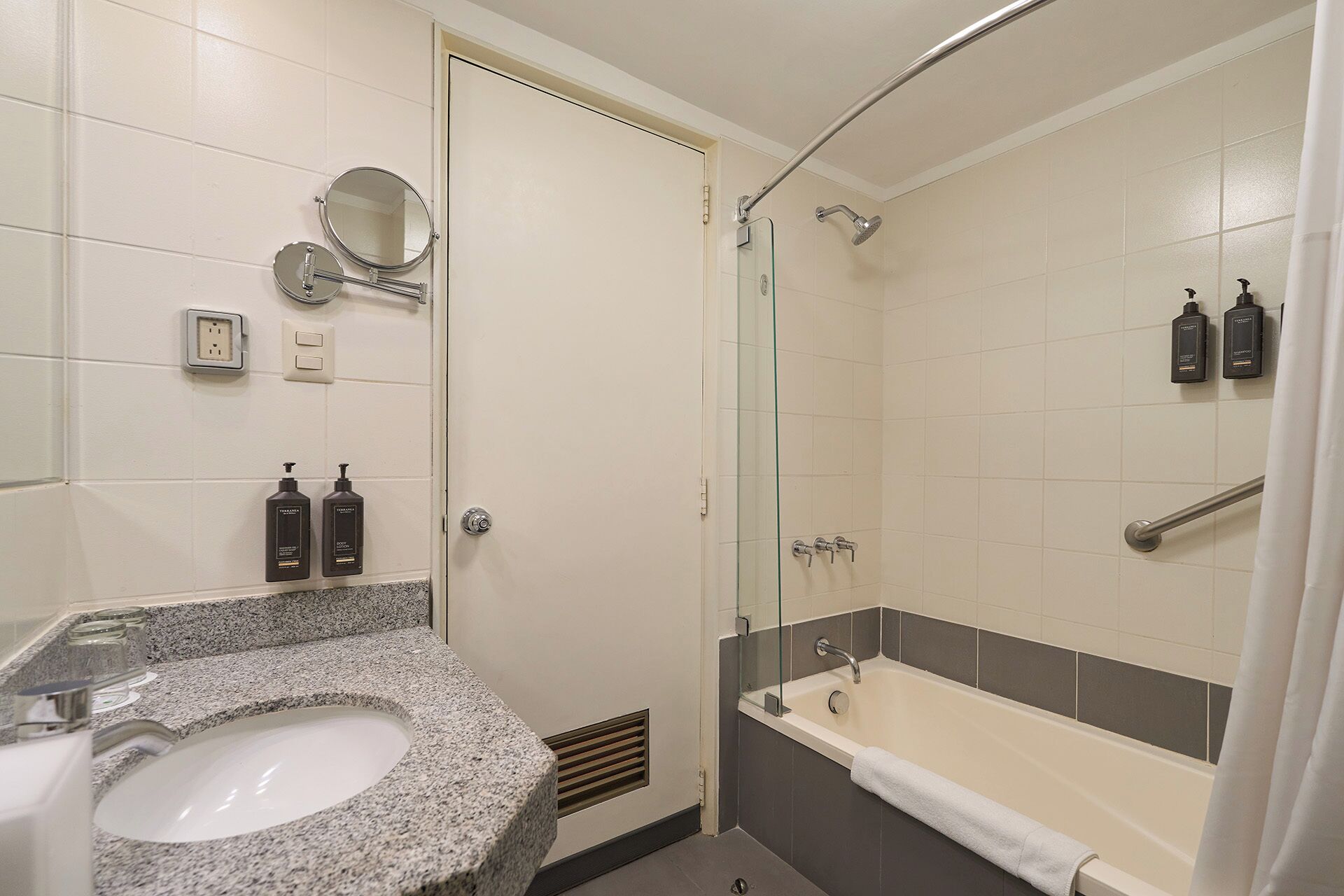 Un baño con lavamanos de encimera de granito, ducha con mampara de vidrio y dos botellas de champú.